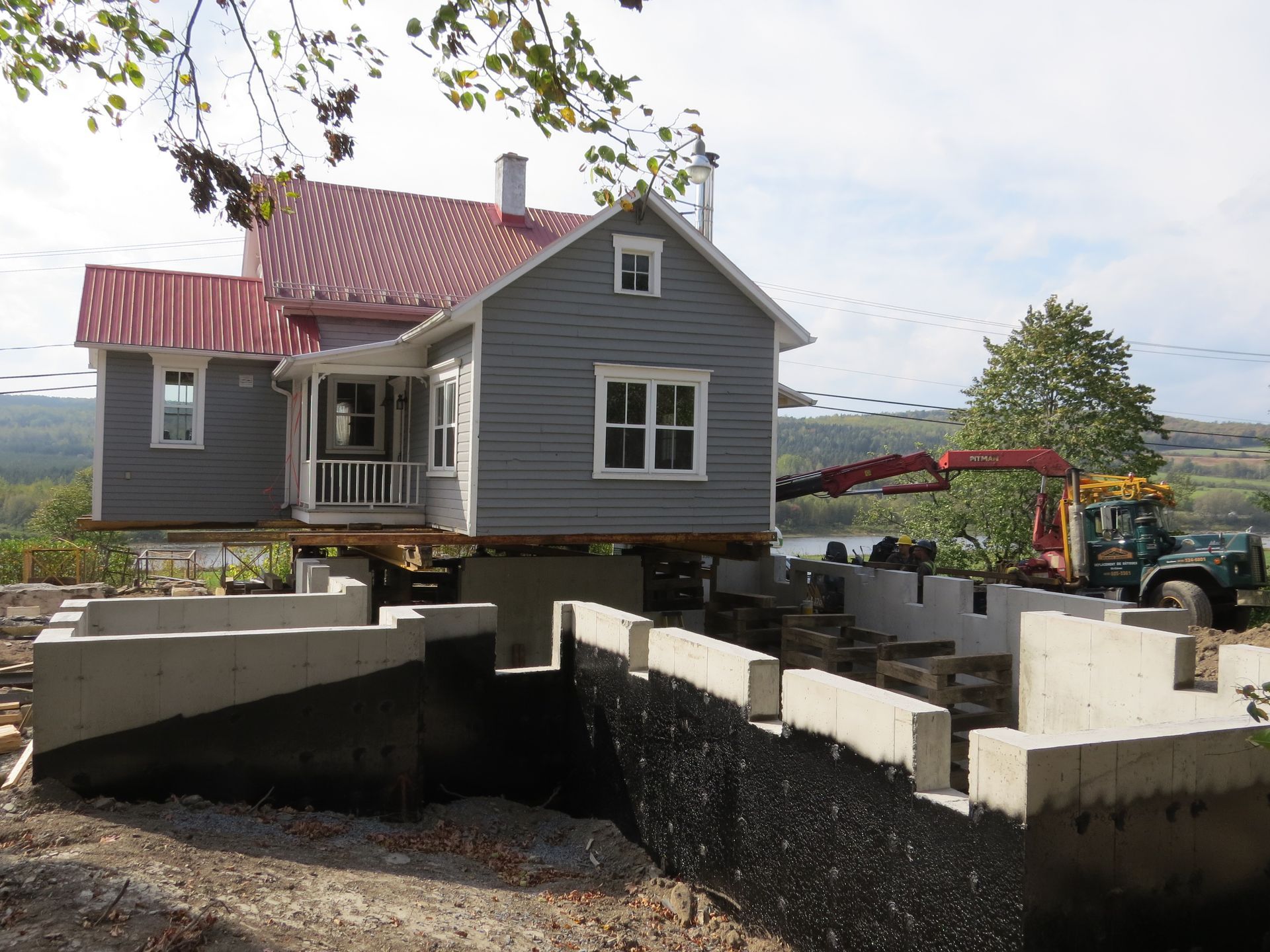 Maison soulevée, nouvelles fondations en construction. Maison grise, toit rouge, vue extérieure. Grue en arrière-plan.