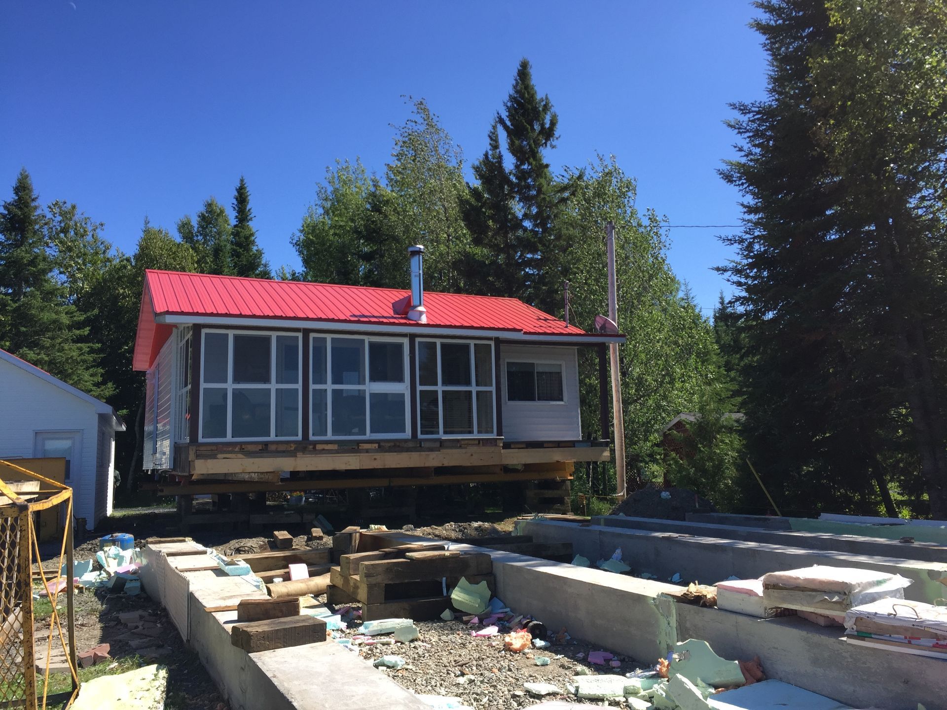 Une cabane au toit rouge est déplacée sur des fondations en béton, entourée d'arbres sous un ciel bleu.