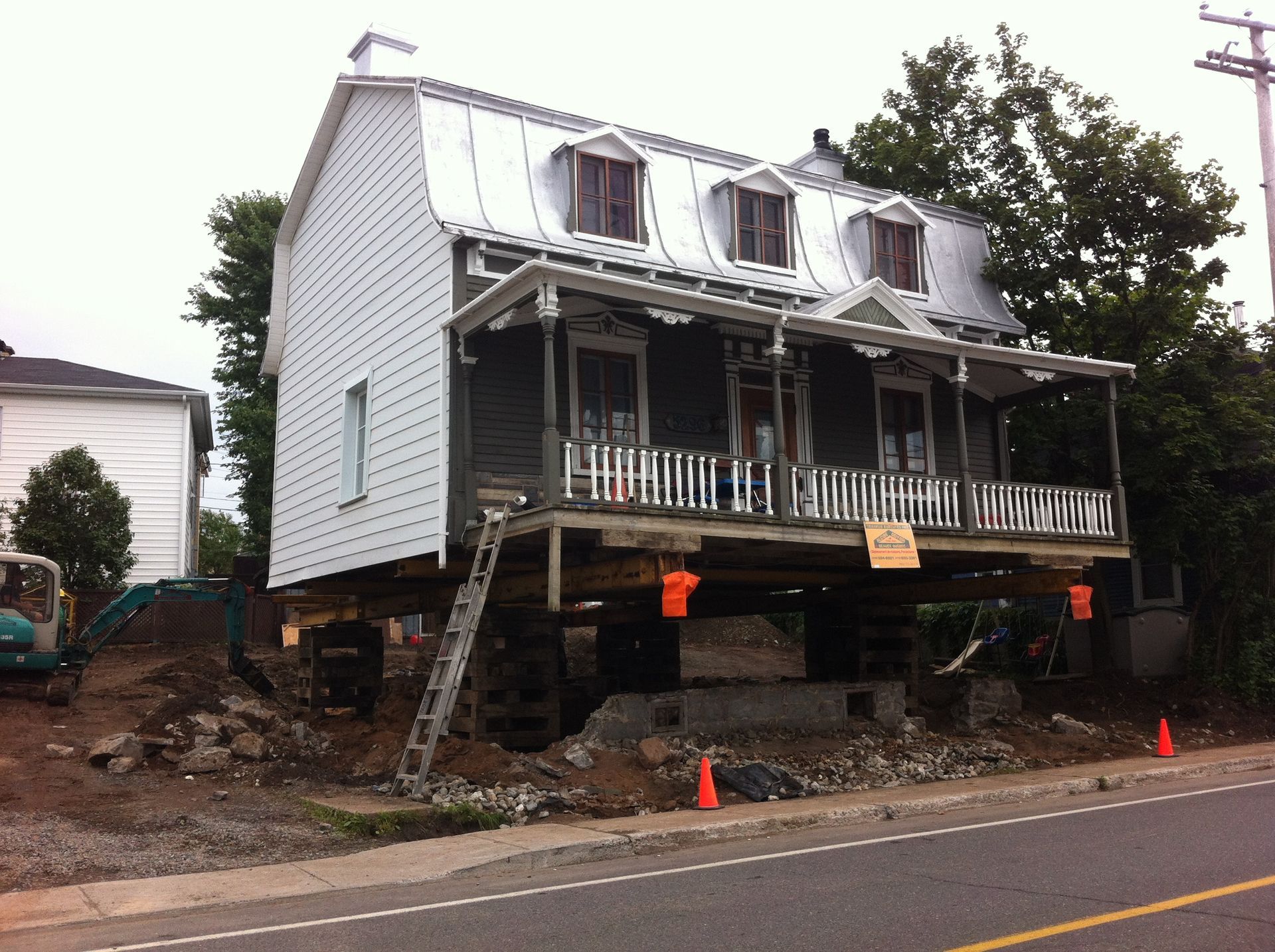 Maison sur pilotis en construction ; terre, route et arbres visibles.