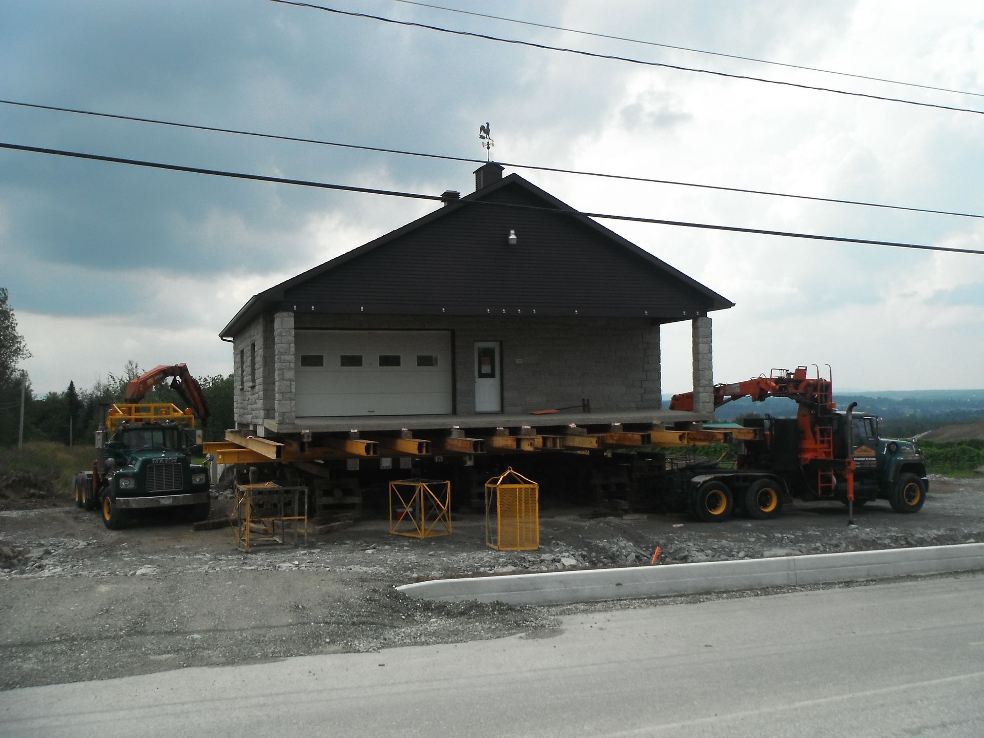 Une maison sur une remorque est transportée sur un chemin de gravier par un camion et des engins de chantier.