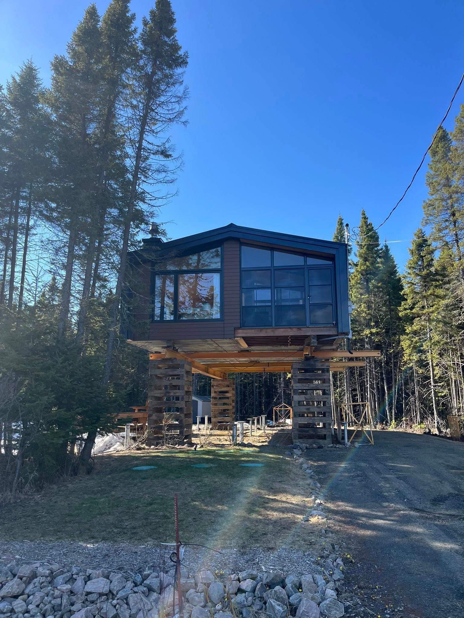 Cabane moderne sur pilotis en bois surélevés, dans une zone boisée sous un ciel bleu.