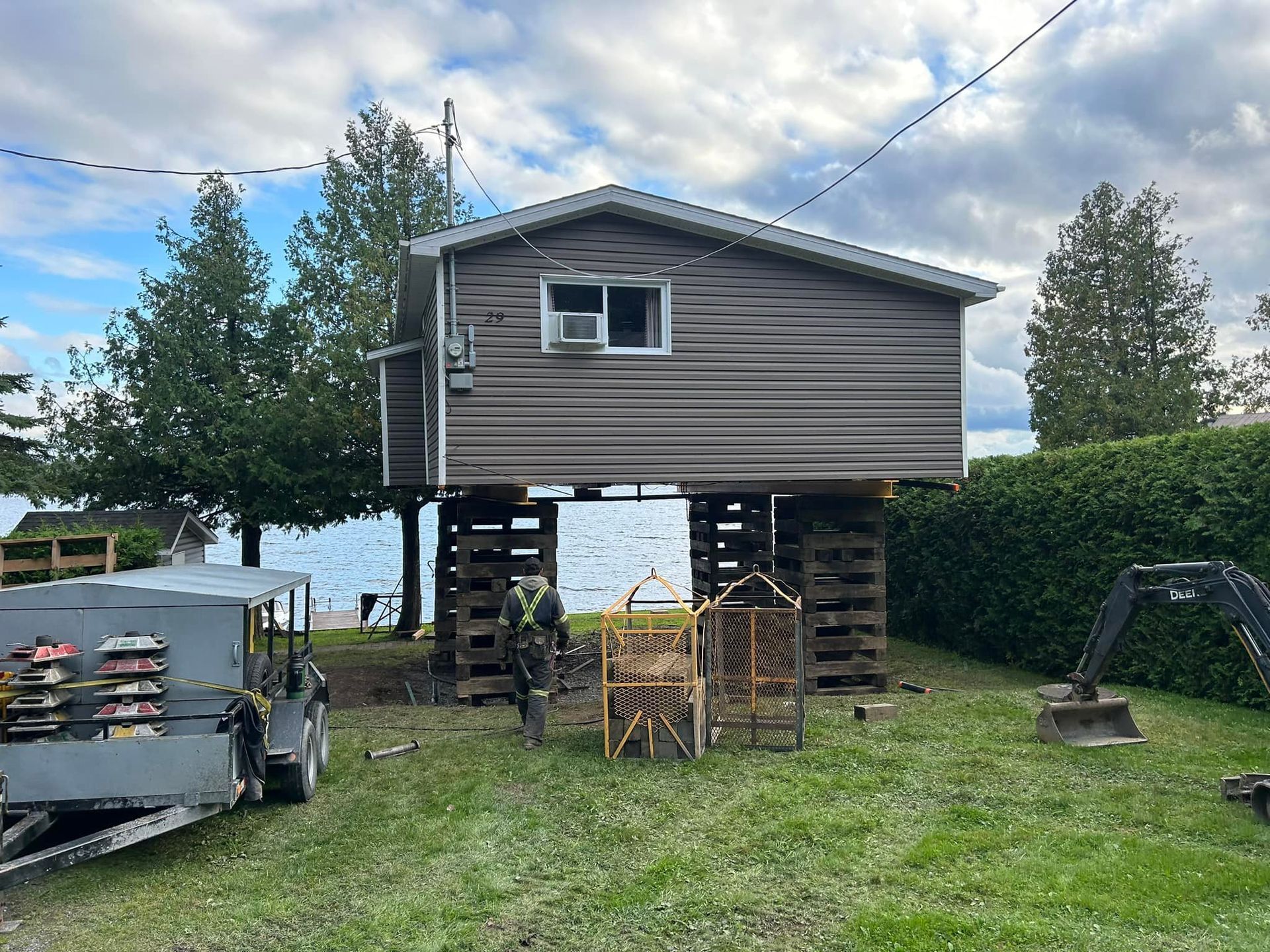 Maison surélevée sur des blocs de bois, près d'un lac. Travaux en cours, ciel nuageux.