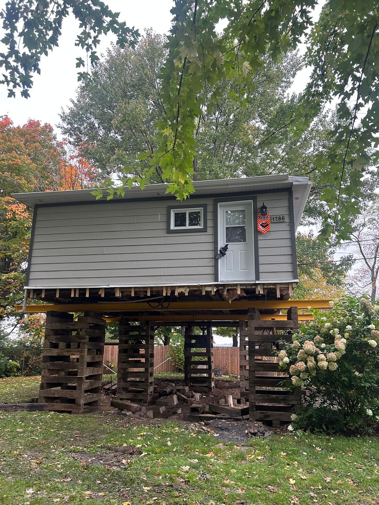 Une petite maison sur pilotis. Bardage gris, porte blanche et petite fenêtre. Dehors, en automne.