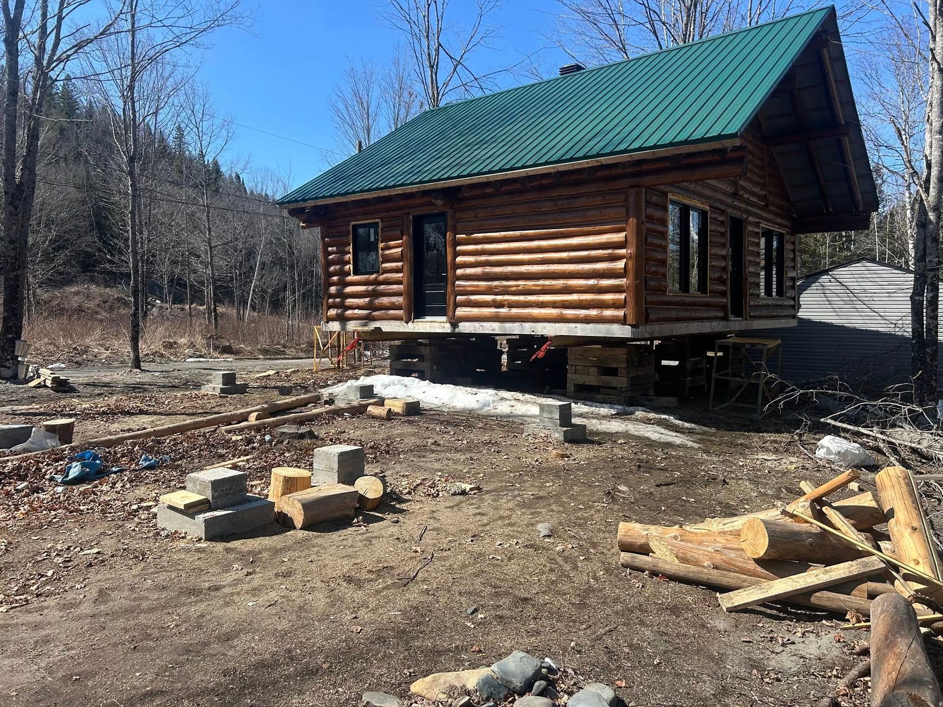 Cabane en rondins avec toit végétalisé sur fondations surélevées, entourée d'arbres et de matériaux de construction.