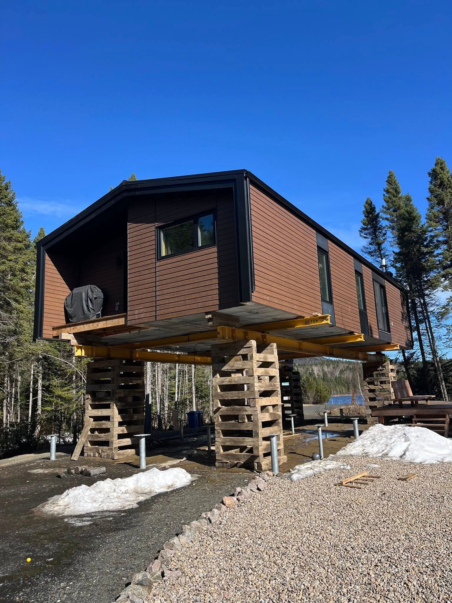 Cabane brune sur pilotis en bois, sous un ciel bleu, entourée d'arbres et de gravier.