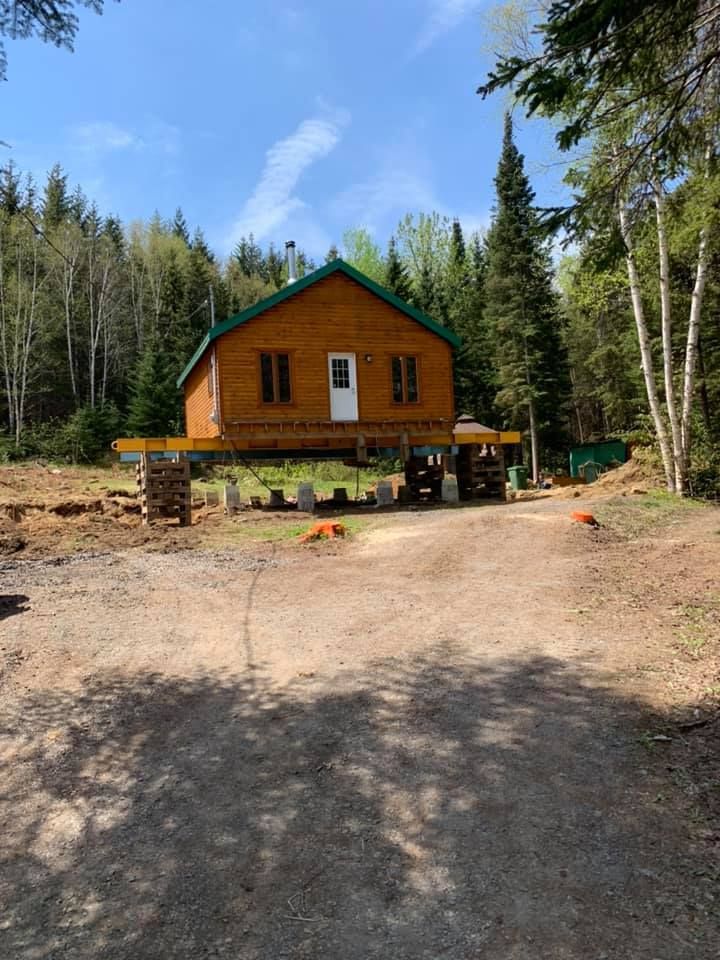 Cabane en bois surélevée sur pilotis dans une clairière en forêt ; chemin de gravier au premier plan.