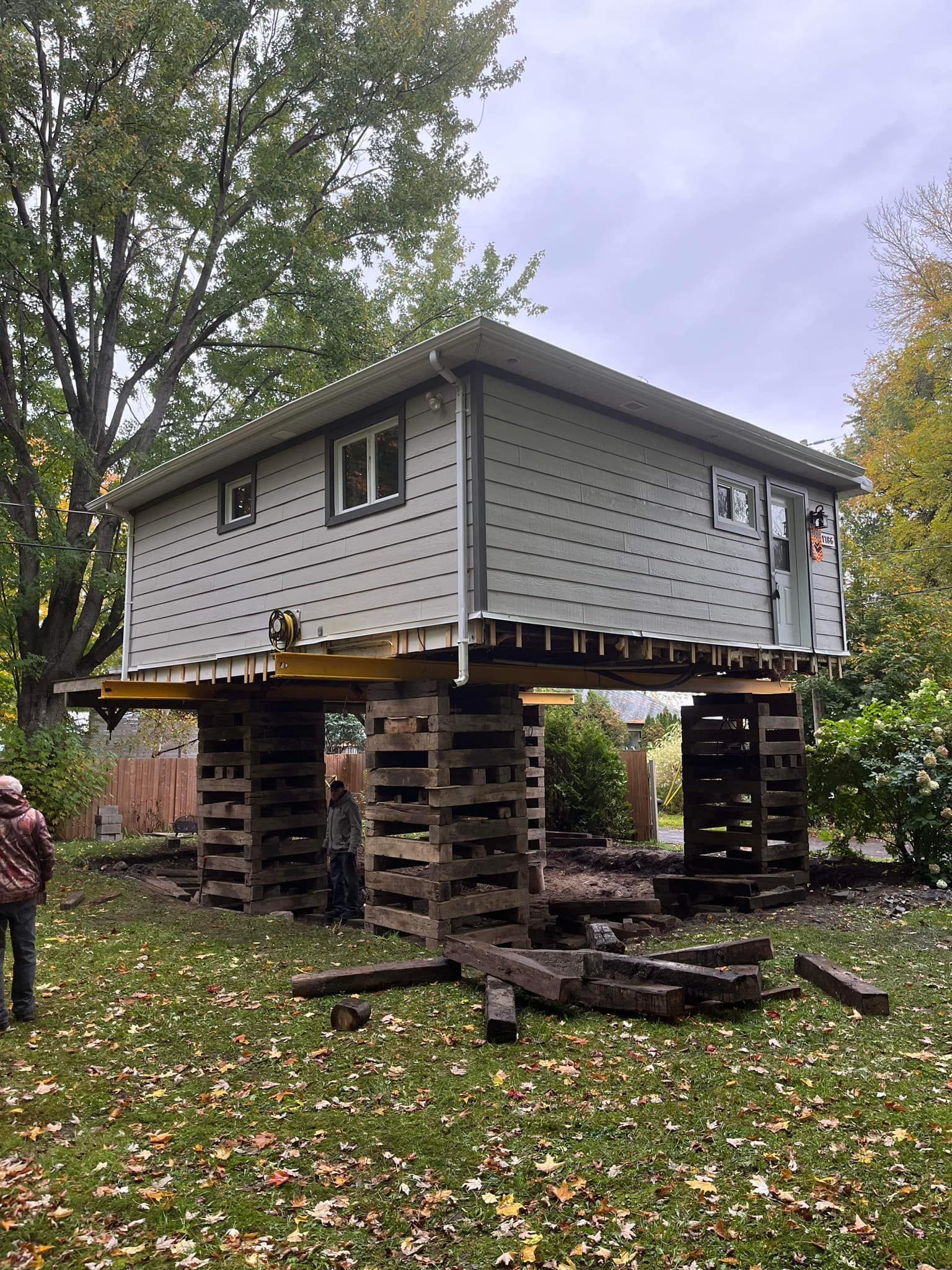 Maison sur pilotis en bois, en cours de levage. Ouvriers à proximité. Herbe verte et arbres en arrière-plan.