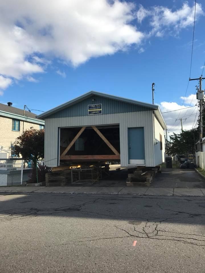 Un bâtiment blanc sur pilotis en bois se dresse au bord d'une route. Un grand triangle en bois est visible à l'entrée.