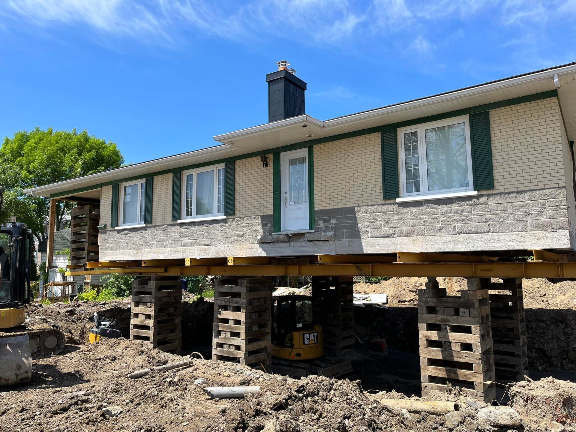 Maison surélevée sur des supports en bois pendant les travaux, entourée de terre et d'engins de chantier.
