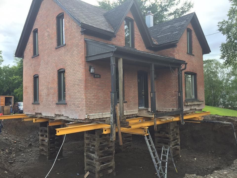 Maison en briques rouges surélevée sur des poutres métalliques jaunes, probablement pour des travaux de fondation.