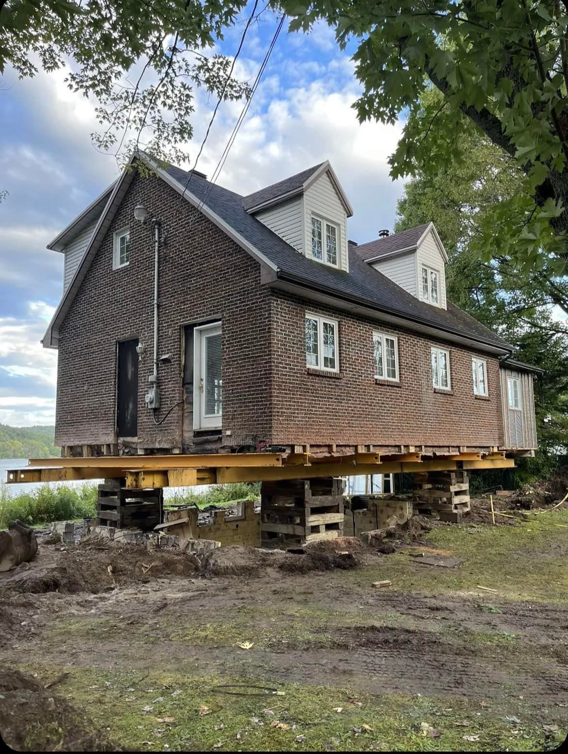 Maison surélevée sur pilotis en bois ; brique et bardage, deux étages, lac en arrière-plan.