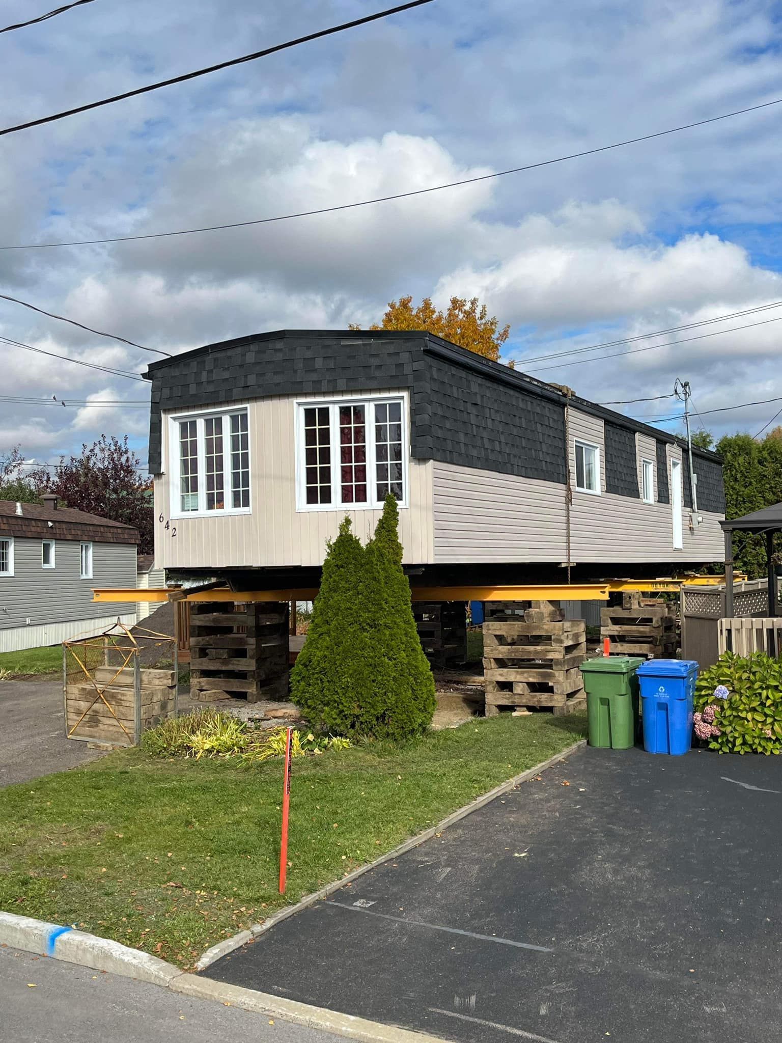 Maison mobile surélevée sur des supports en bois, sur un terrain herbeux, avec des poteaux électriques.