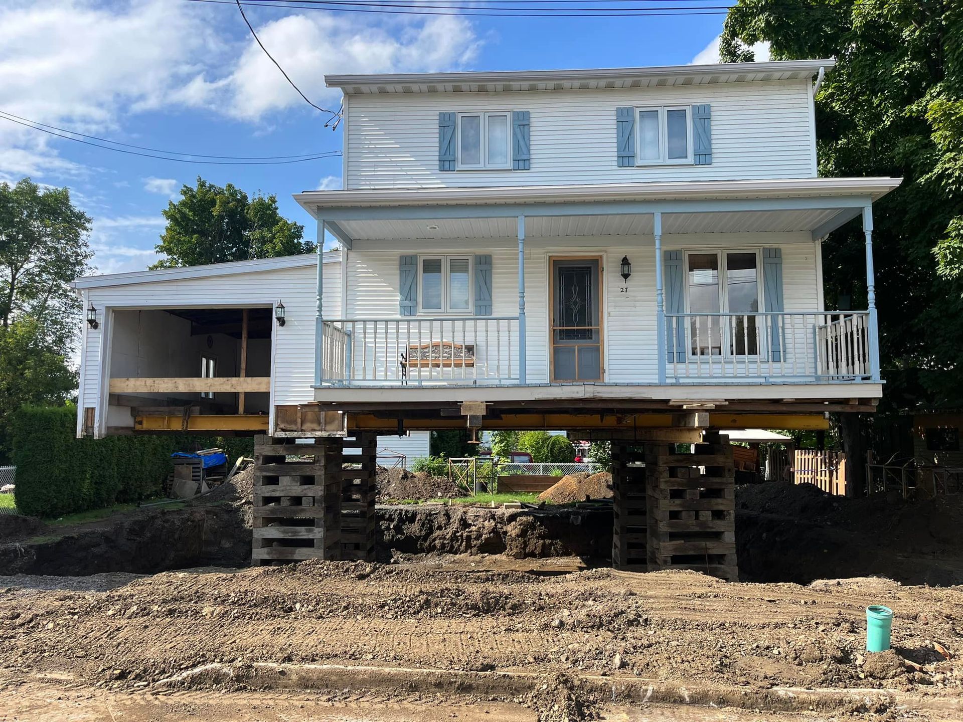 Une maison blanche à deux étages est surélevée sur des supports en bois au-dessus d'un chantier de construction.