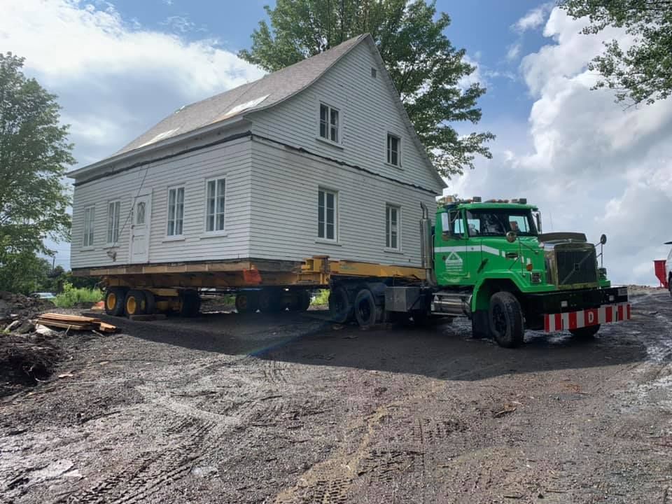 Une maison sur remorque déplacée par un camion vert sur un chemin de terre.