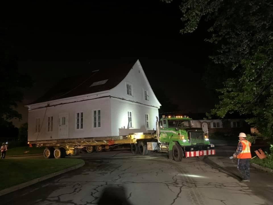 La Maison Blanche, transportée de nuit sur un camion plateau par un camion vert, est supervisée par des ouvriers en gilets.