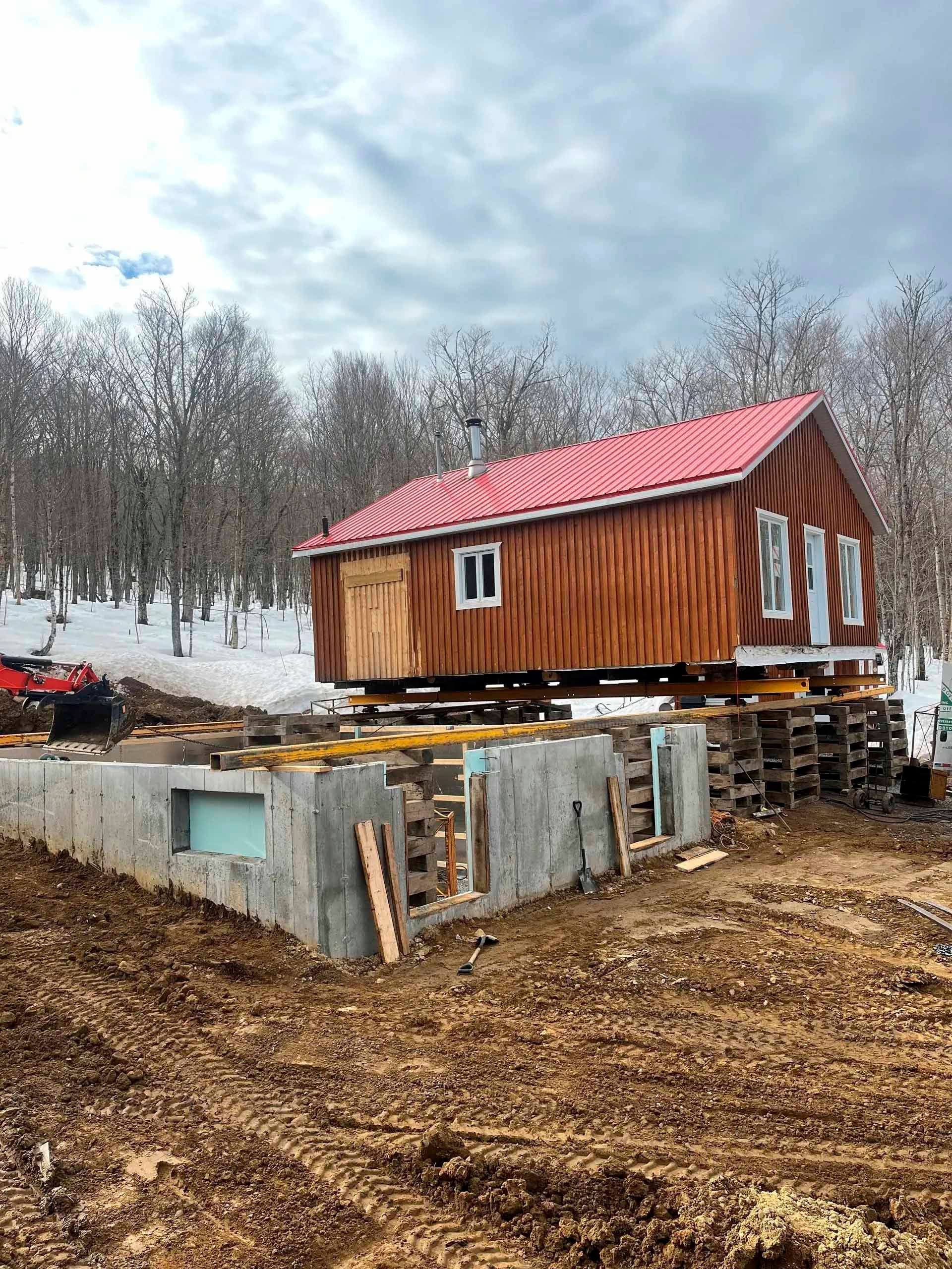 Maison sur parpaings, en cours de déplacement sur des fondations en béton, terre et arbres dénudés en arrière-plan.