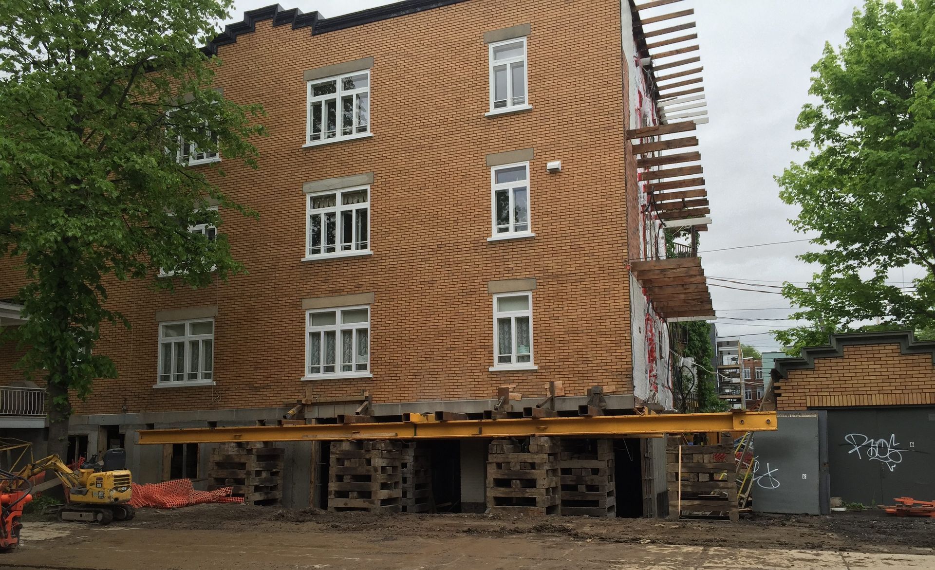 Bâtiment surélevé sur des supports en bois pendant sa construction. Façade en briques, fenêtres blanches et poutres jaunes.