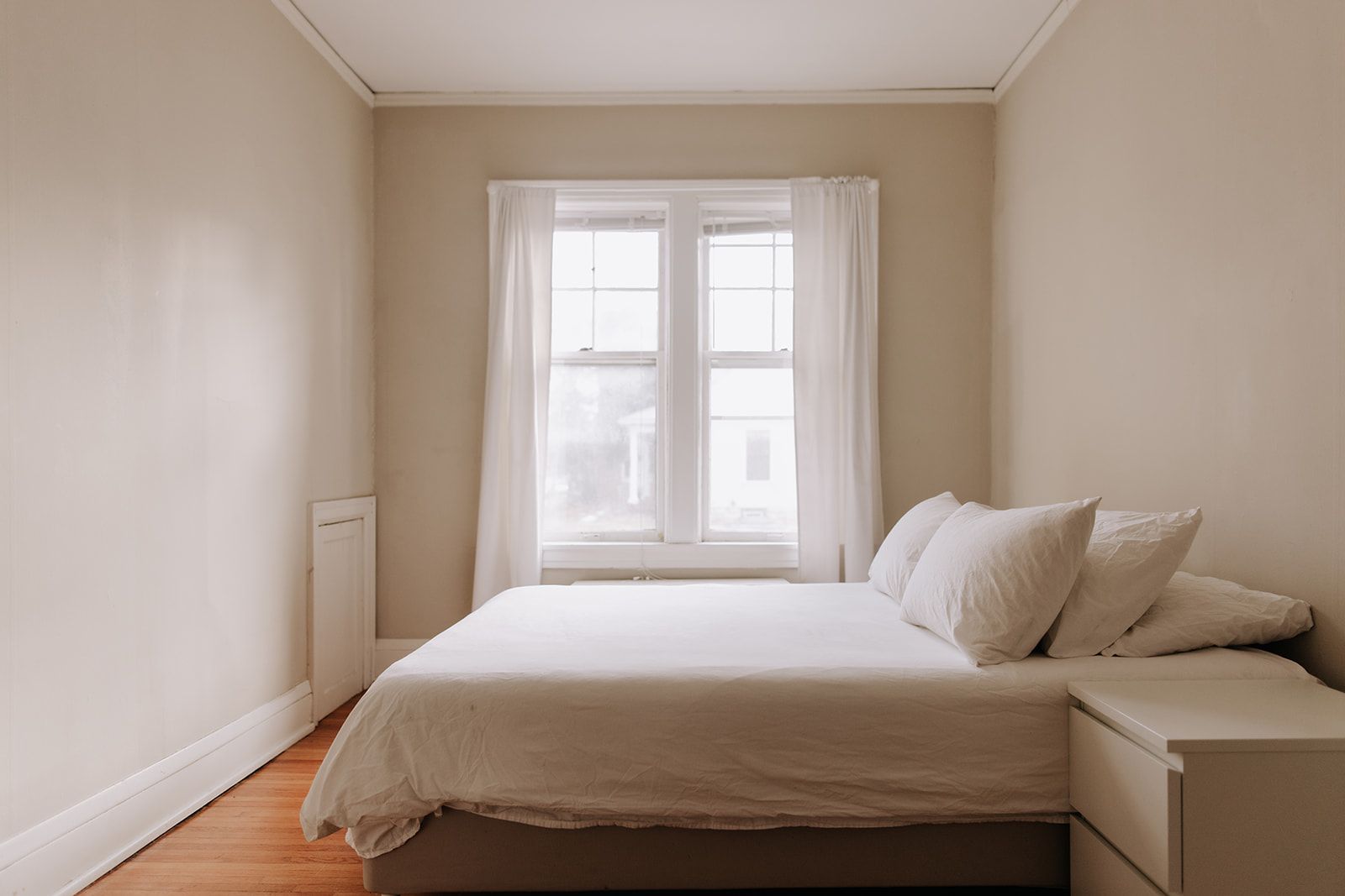 A simple apartment bedroom with warm wood floors. The bed has all white linens. The large window has light, white curtains. A small white nightstand sit by the bed. South Minneapolis apartments by RightMove Properties.
