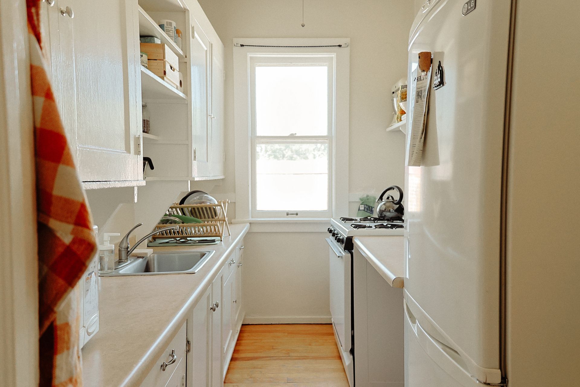 A New York style apartment with white cabinetry and wood floors. Features a window at the back of the room and shelving over the sink and oven. Apartments in Linden Hills by RightMove Properties. 