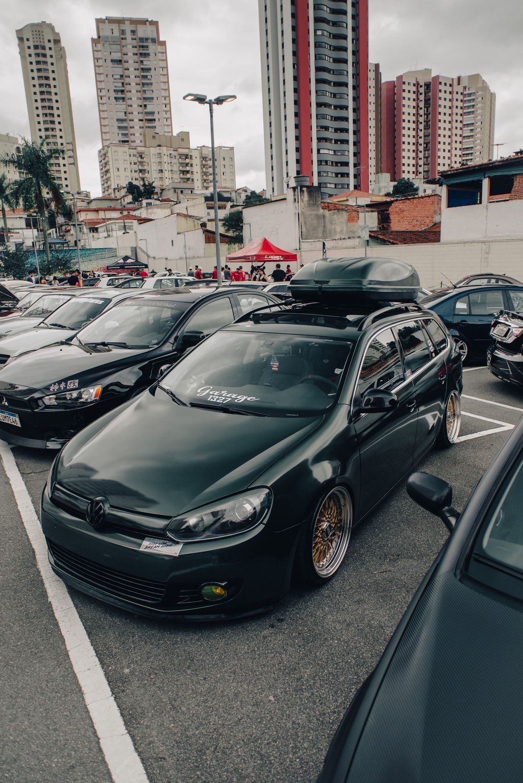 Dark green station wagon parked at a car show, roof cargo carrier, low profile tires, urban setting with tall buildings.