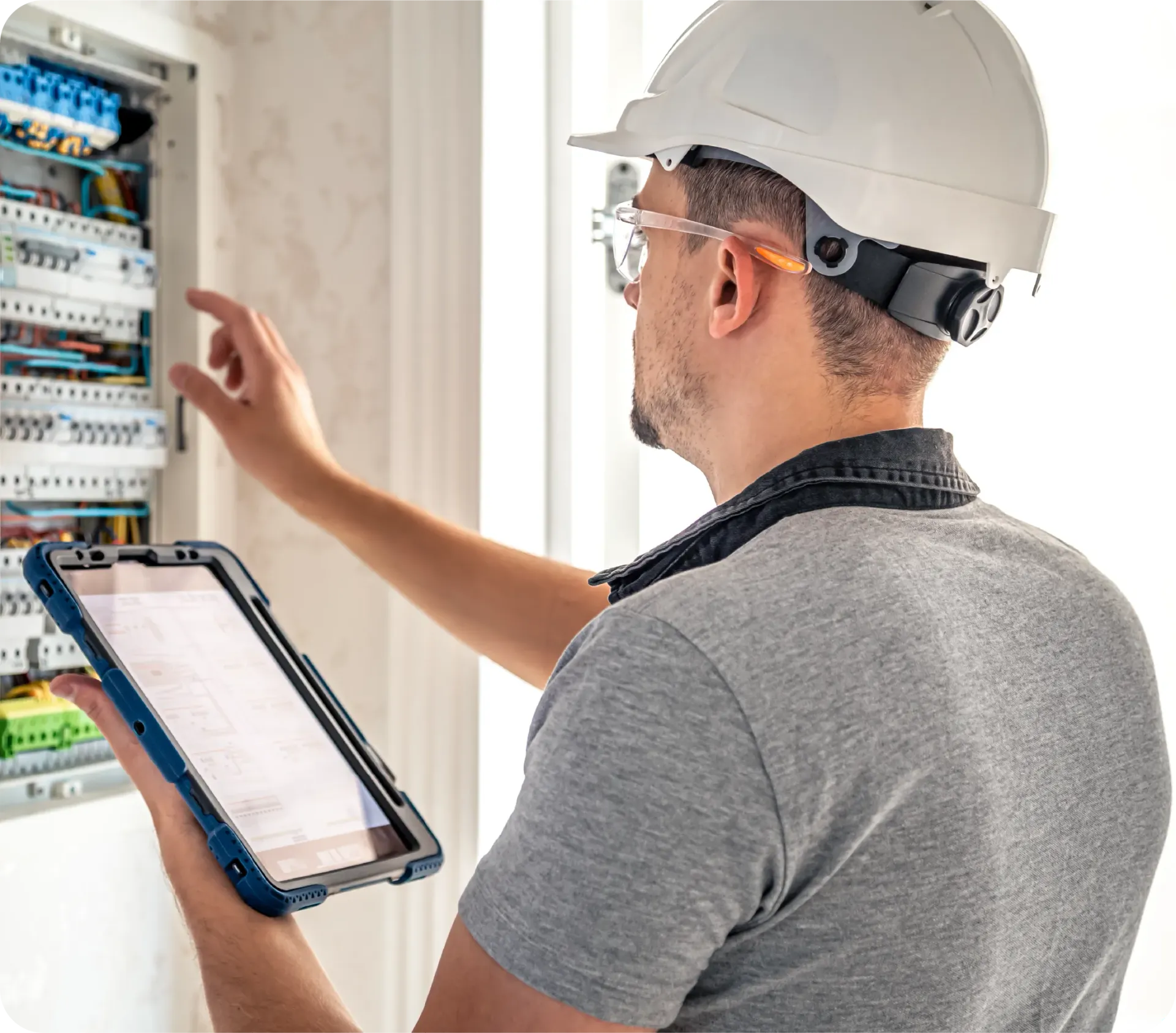 Electrician inspecting electrical panel, holding tablet, wearing hard hat and safety glasses.