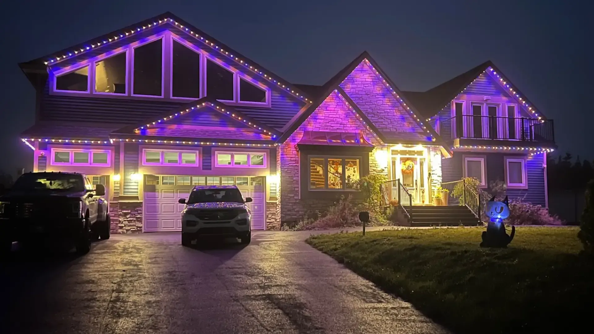 House at night, illuminated with purple and white lights. Cars parked in the driveway.