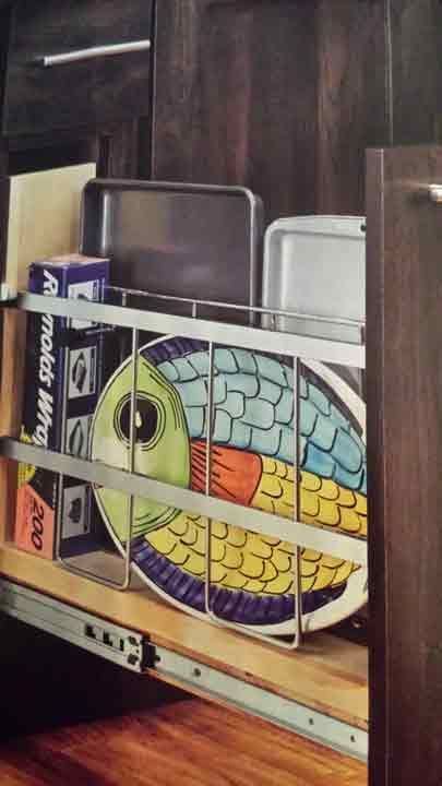 Pull-out cabinet with a decorative fish platter, baking pans, and food wrap, on a wooden shelf.
