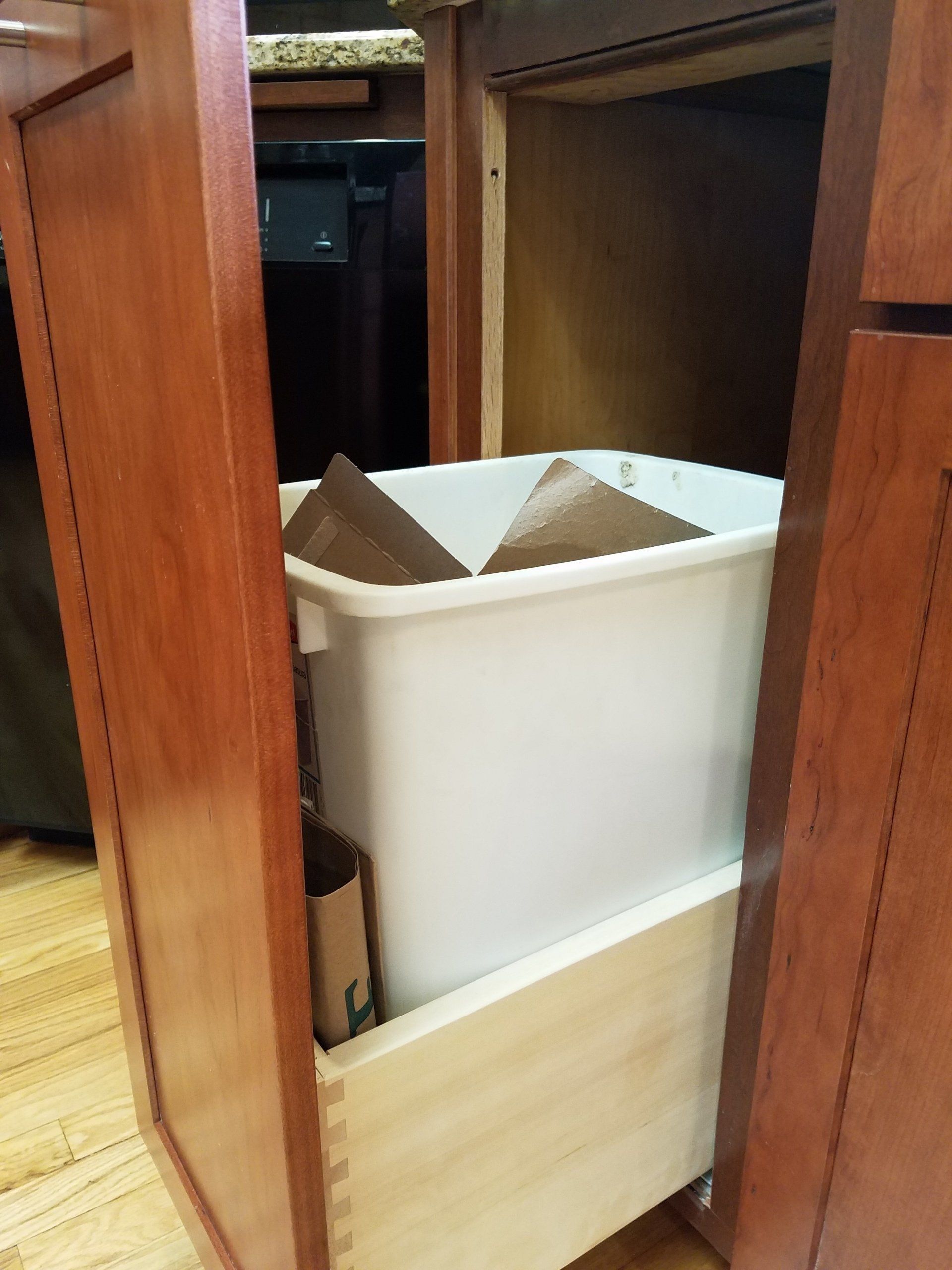 Pull-out kitchen trash bin. White bin sits on wooden drawer, inside a cherry-wood cabinet.