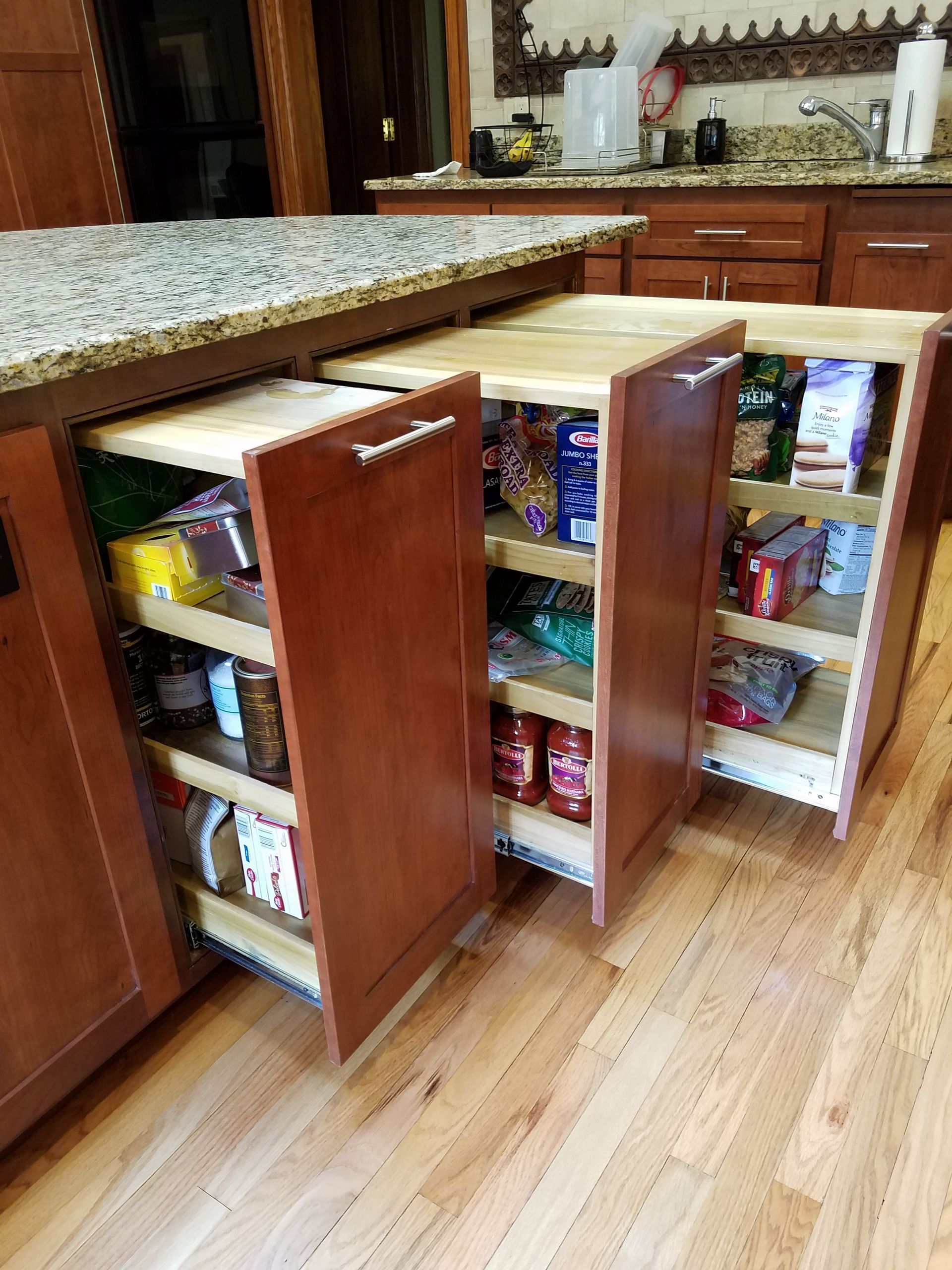 Pull-out pantry cabinets in a kitchen island, filled with food items. Wooden cabinets, granite countertop, and wooden floor.