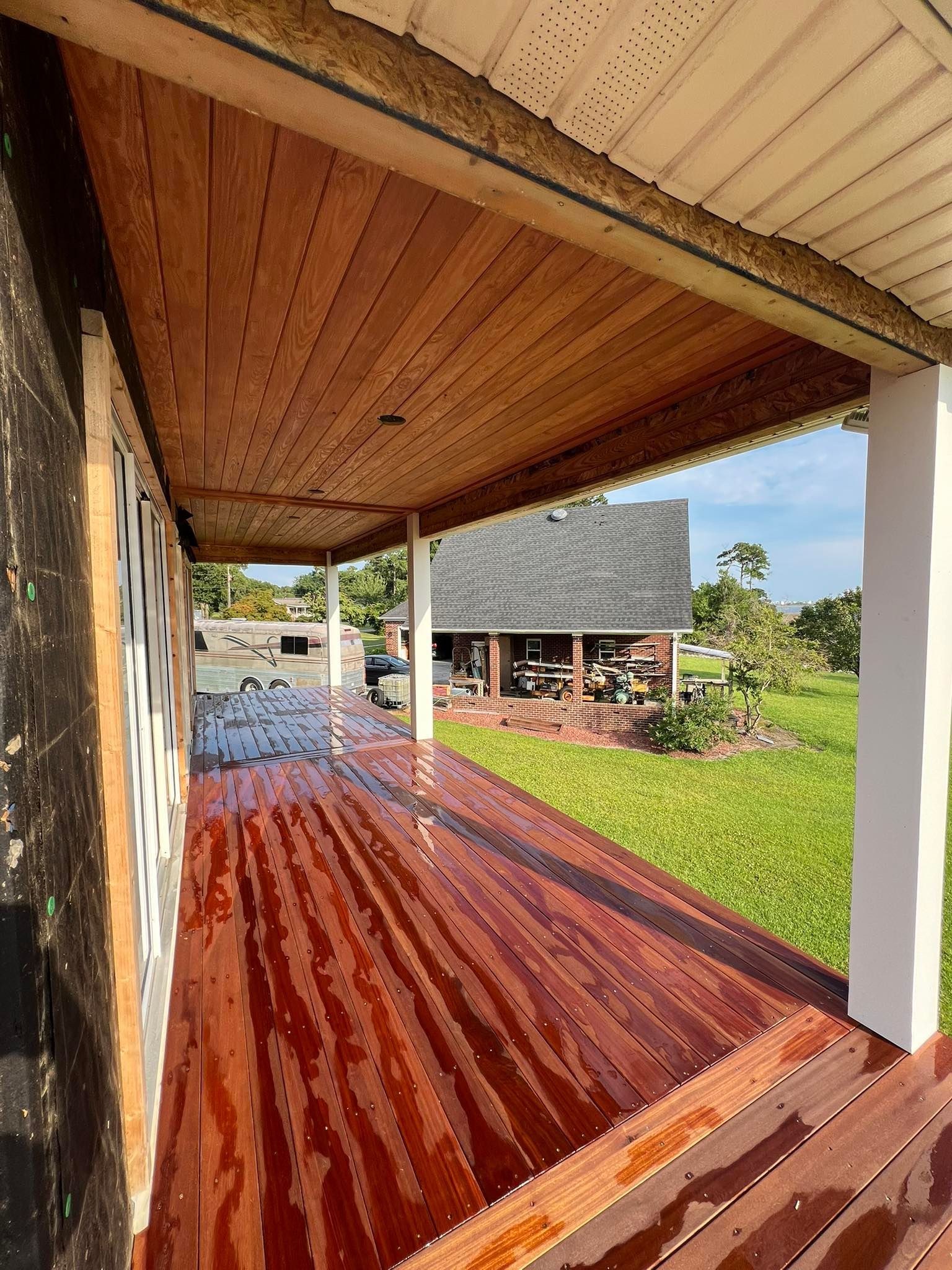 Wooden porch with brown planks, ceiling, and trim. A house and green lawn are in the background.