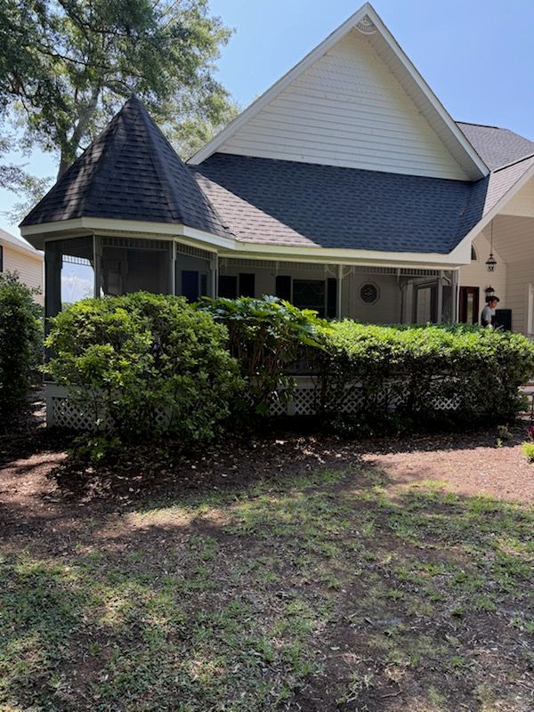 A house with a turret, porch, and green bushes. Gray roof, white trim, and a sunny outdoor setting.