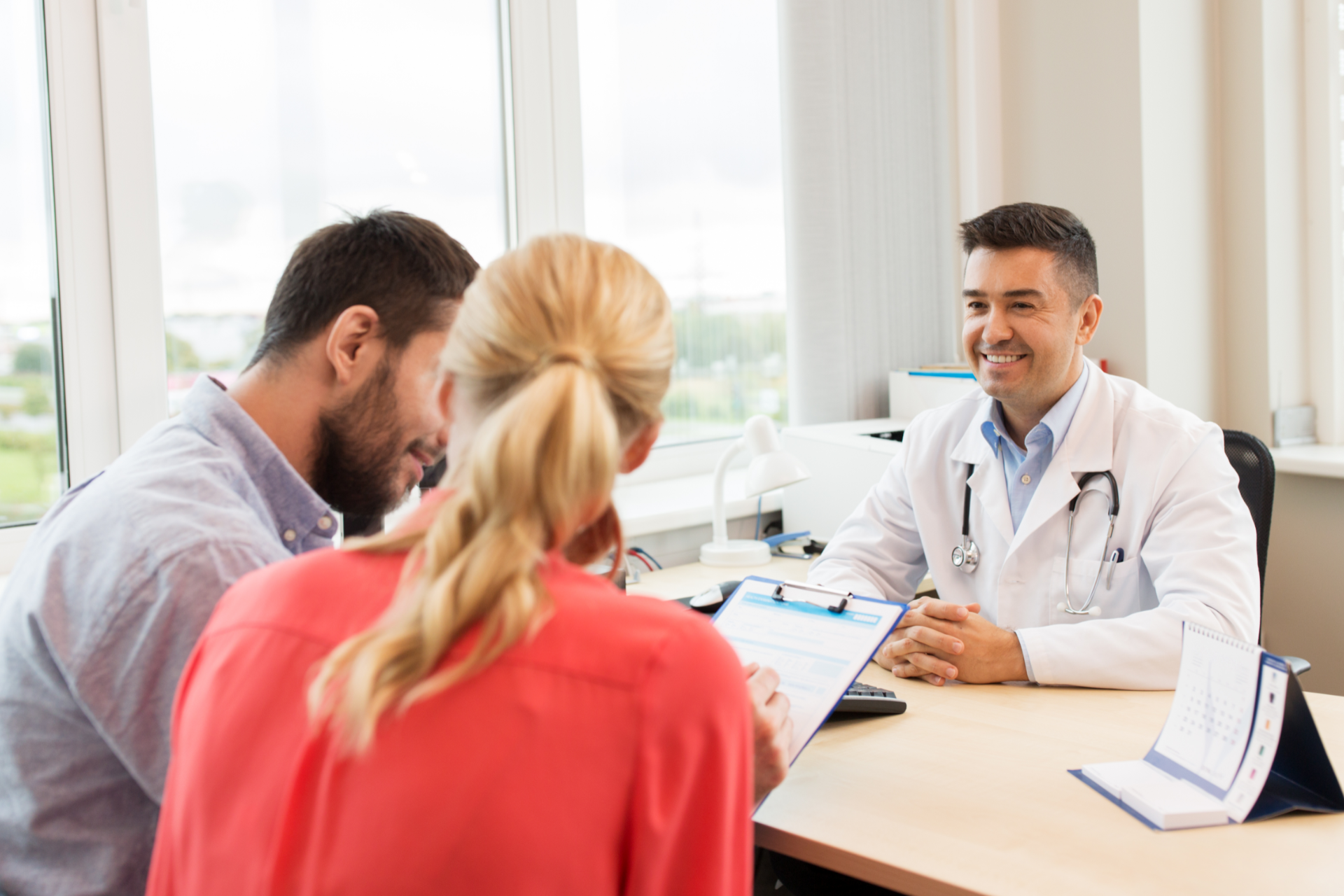 Person in gray blazer holds pen, poised over paperwork at a wooden desk with another person and laptop visible.