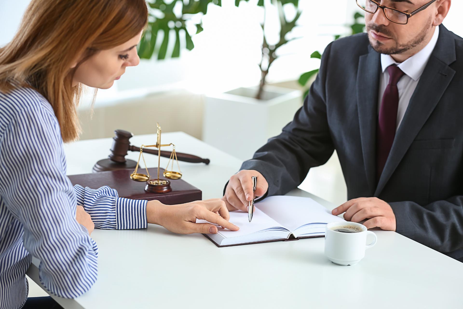 Person in gray blazer holds pen, poised over paperwork at a wooden desk with another person and laptop visible.