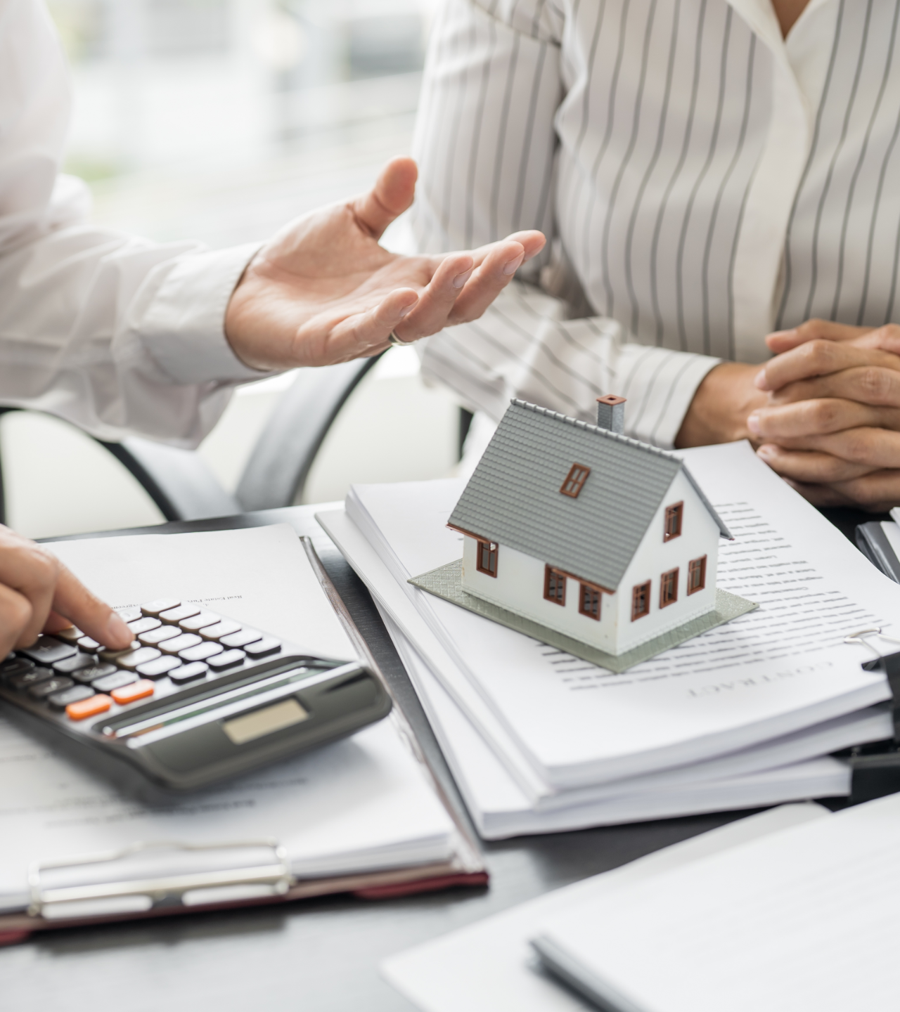 Person in gray blazer holds pen, poised over paperwork at a wooden desk with another person and laptop visible.