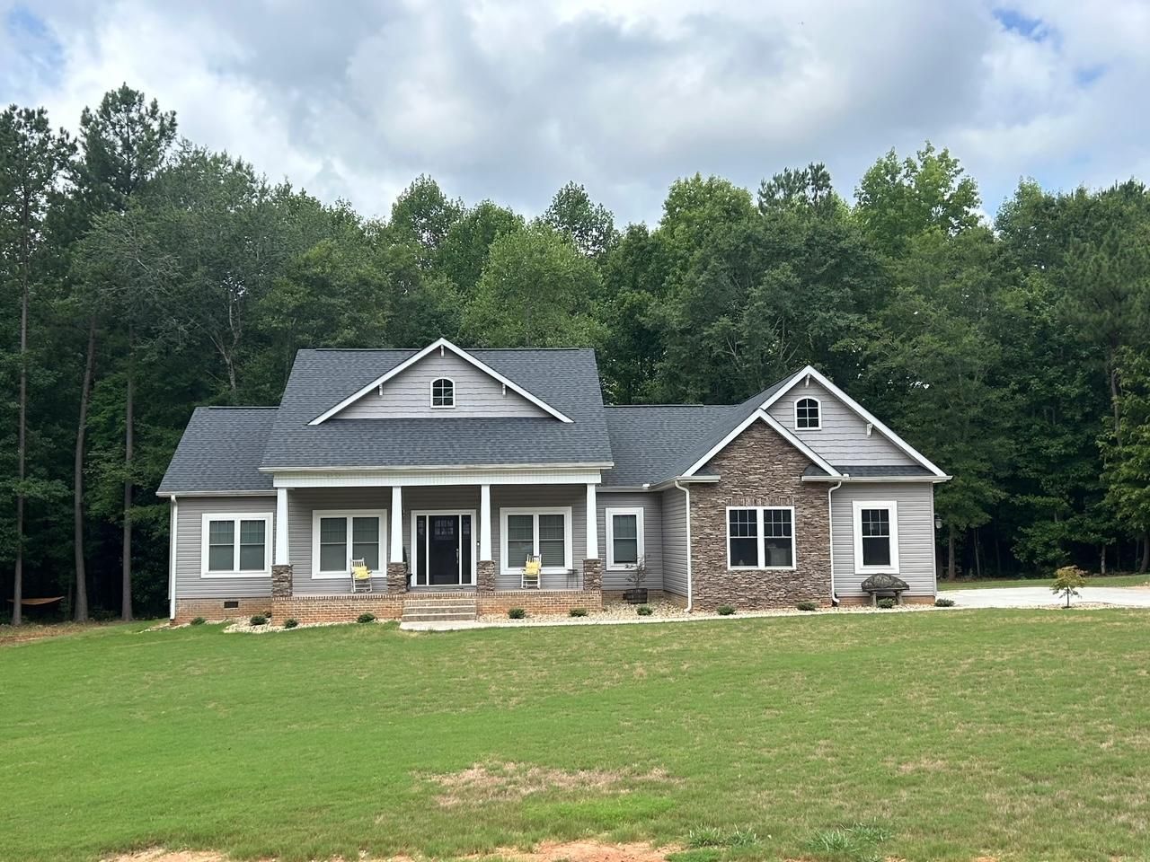 Ranch-style house with gray siding, stone accents, and dark roof, set on a green lawn with a tree line backdrop.