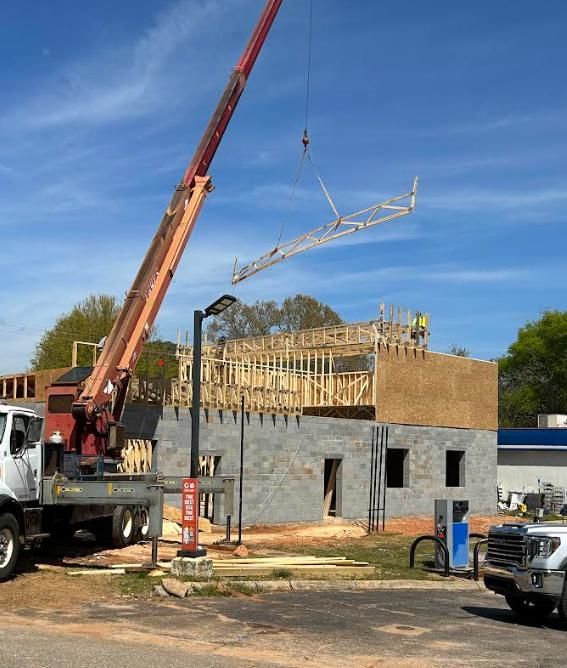 Construction site with crane lifting roof truss; wooden frame structure being built, blue sky.