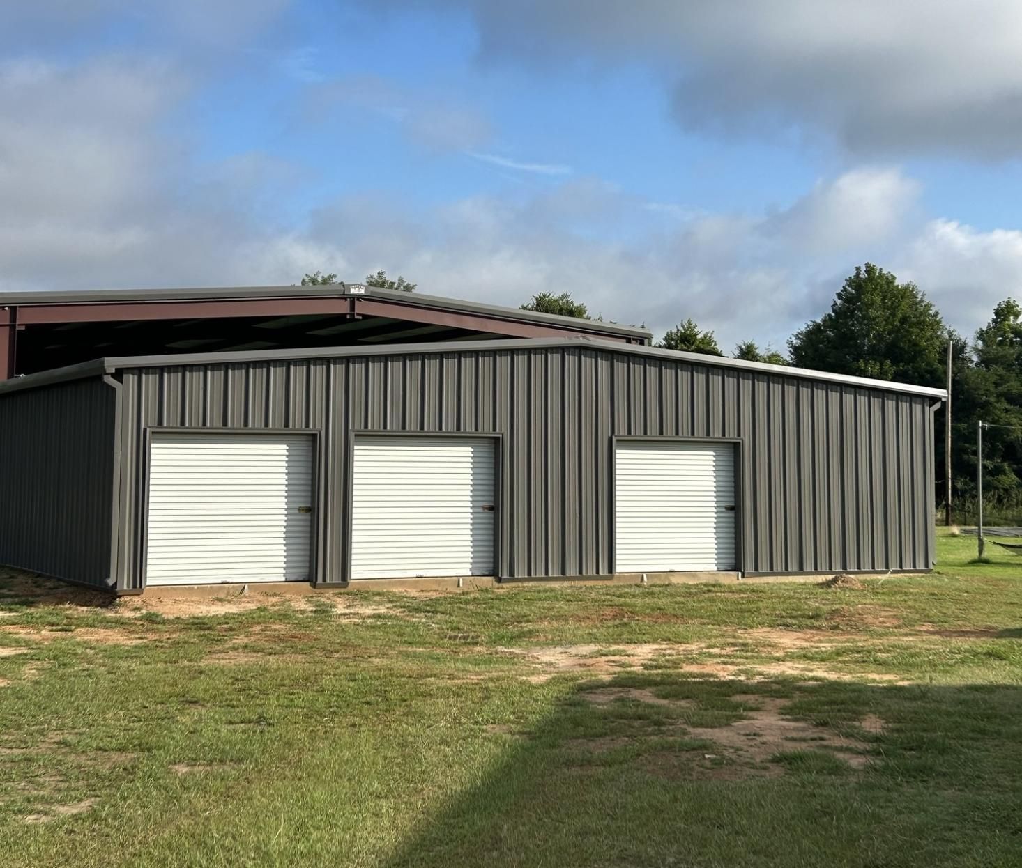 Gray metal building with three white garage doors on a grassy lawn under a partly cloudy sky.