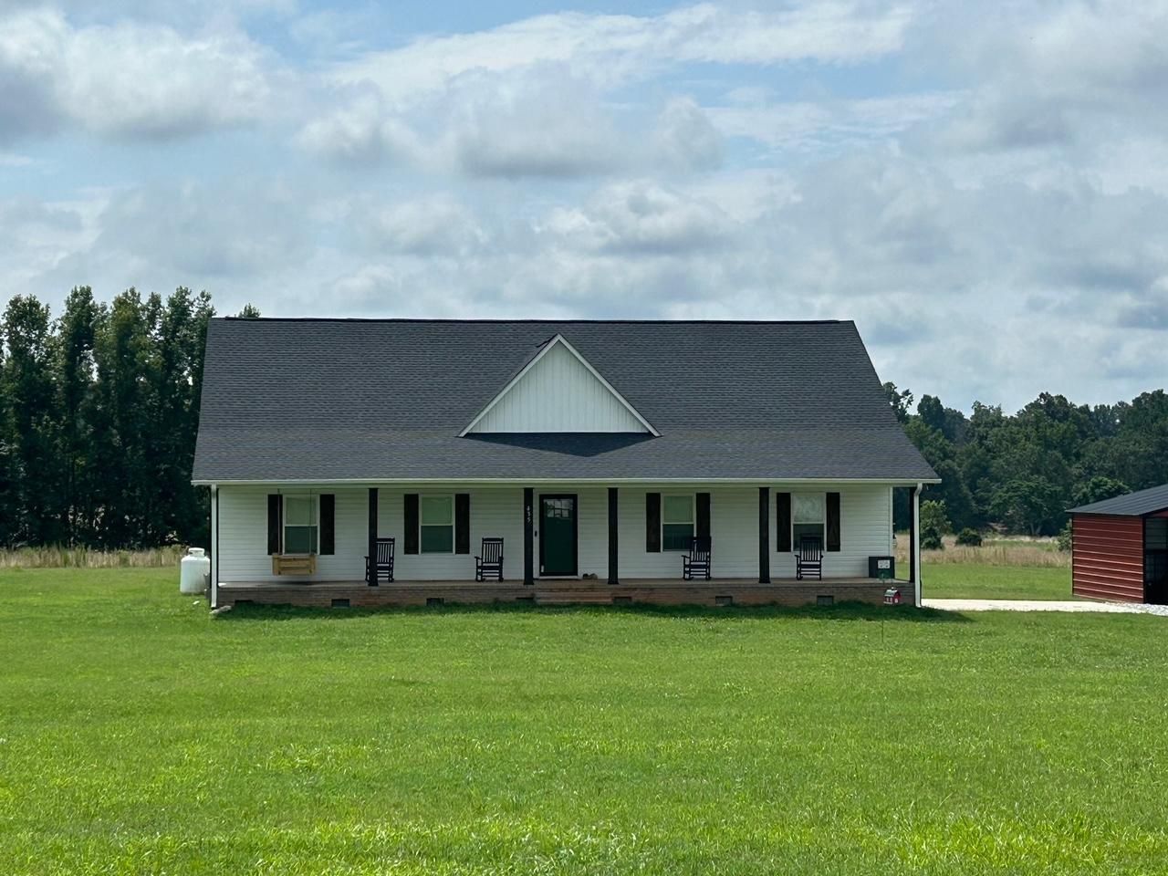 White farmhouse with black shutters and a dark roof, on a grassy field under a cloudy sky.