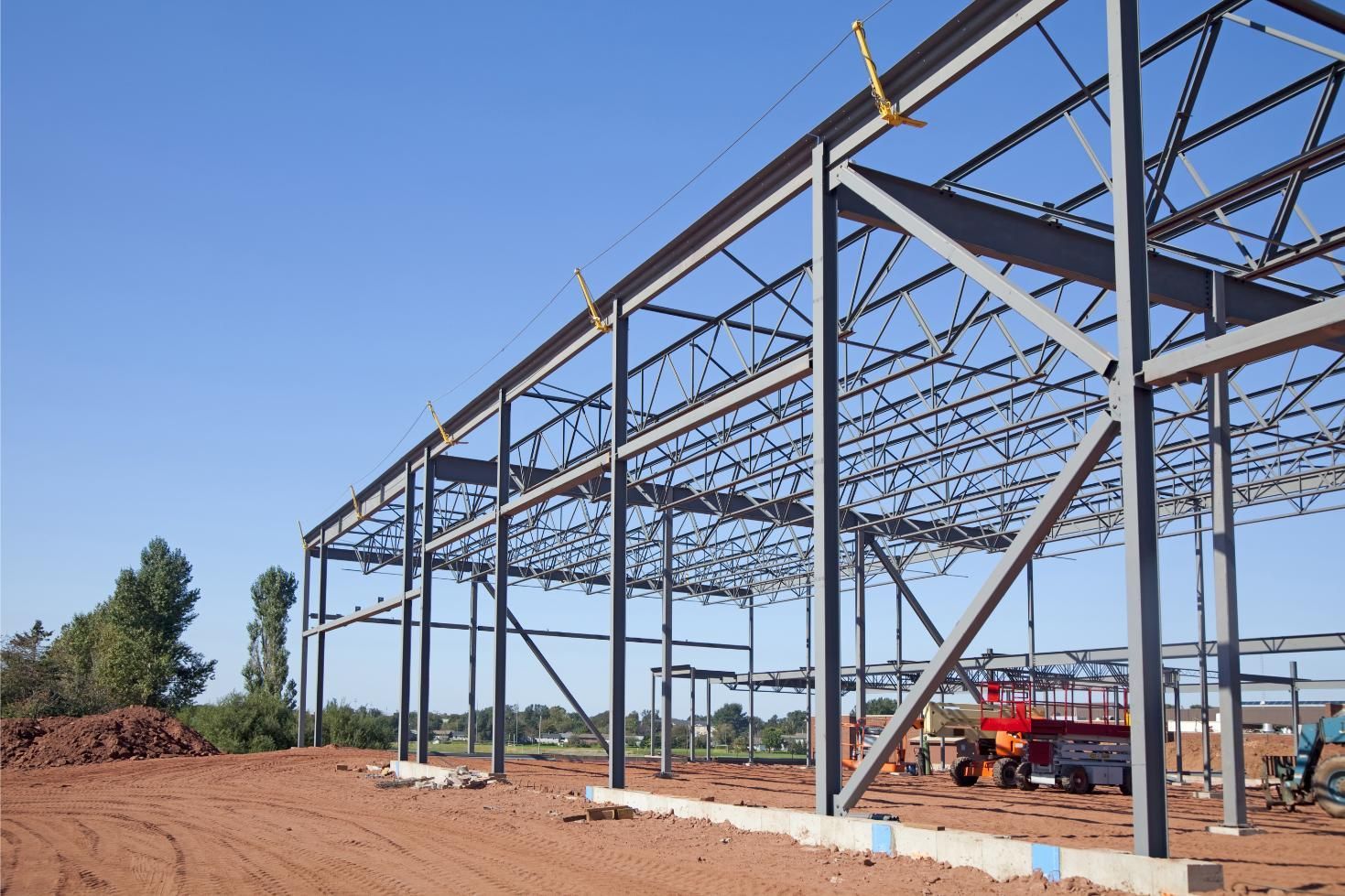 Steel frame of a building under construction on a dirt lot, blue sky background.