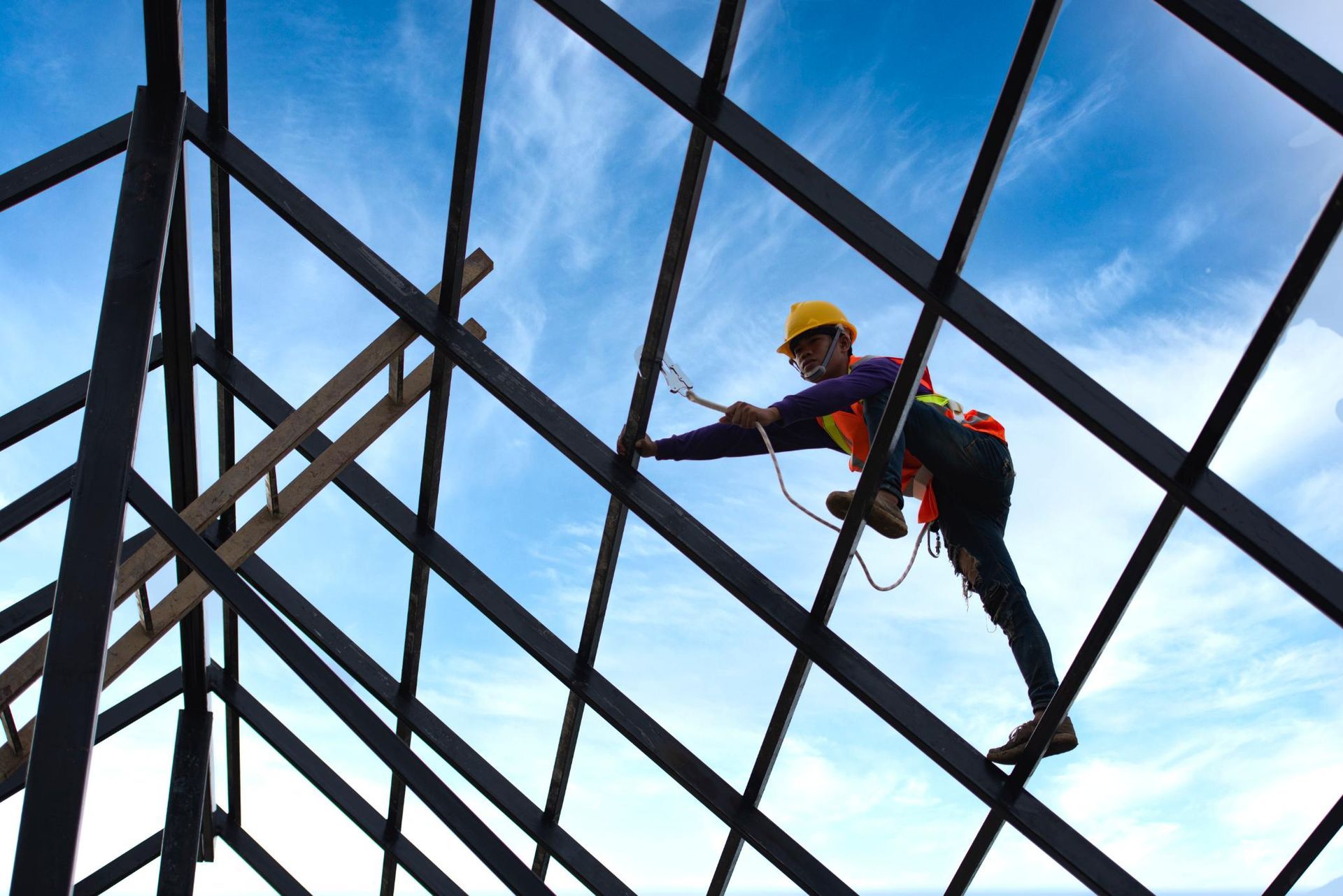 Construction worker on a roof frame, wearing a hard hat and safety harness, against a blue sky.