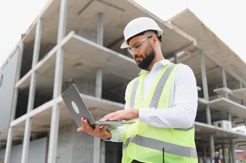 Construction worker in hard hat and vest uses a laptop at a building site.