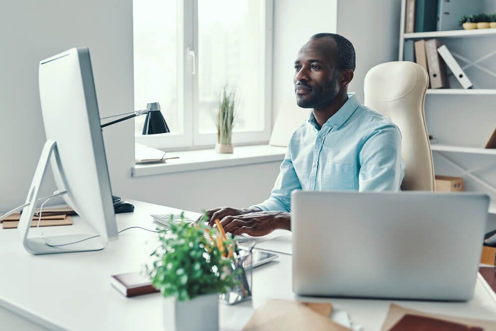 A man is sitting at a desk in front of a computer and a laptop.