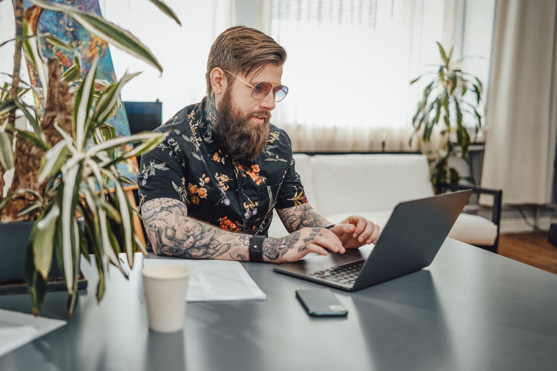 Man with beard and tattooed body wearing stylish clothing doing his work on a laptop in cosy room.