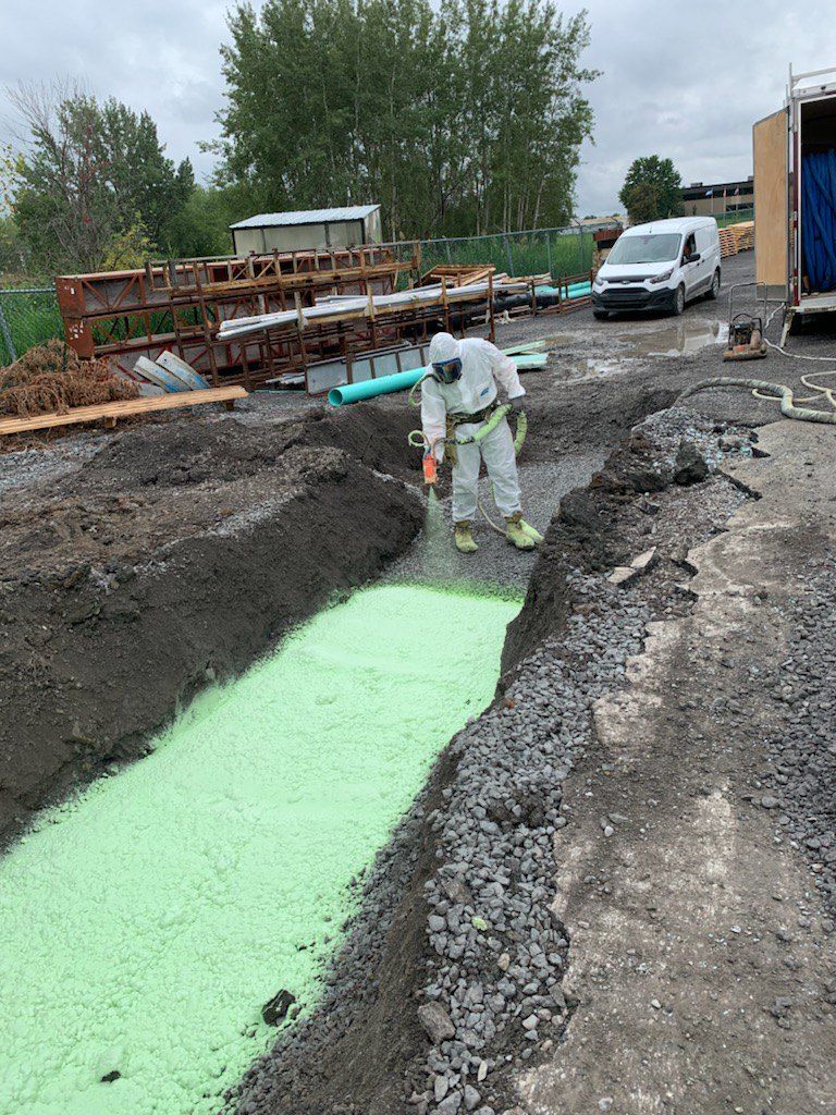 Une personne en combinaison de protection inspecte une tranchée remplie de mousse verte sur un chantier de construction.