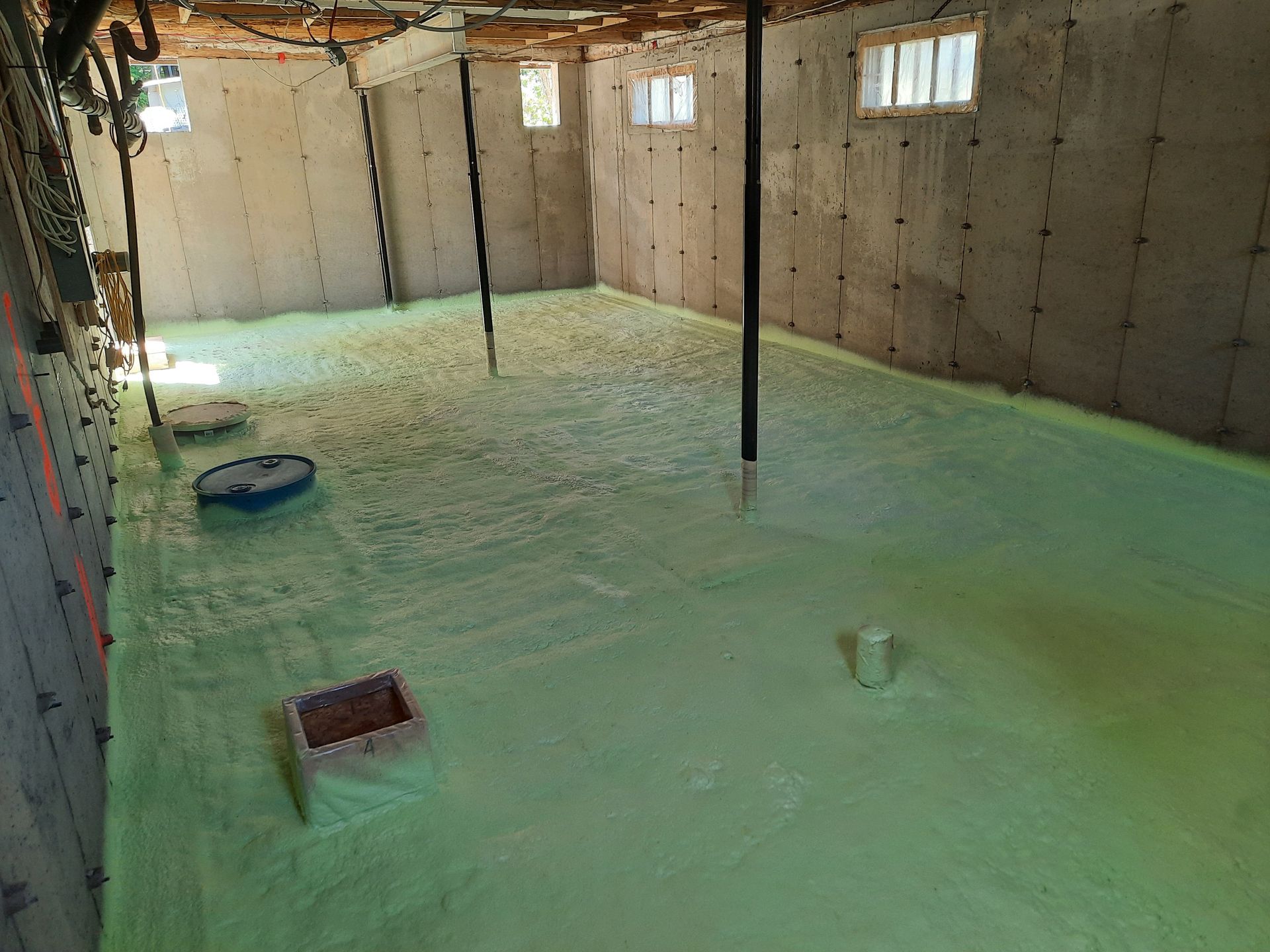 Sous-sol avec isolation en mousse verte au sol. Murs en béton, poutres de soutien et petites fenêtres visibles.