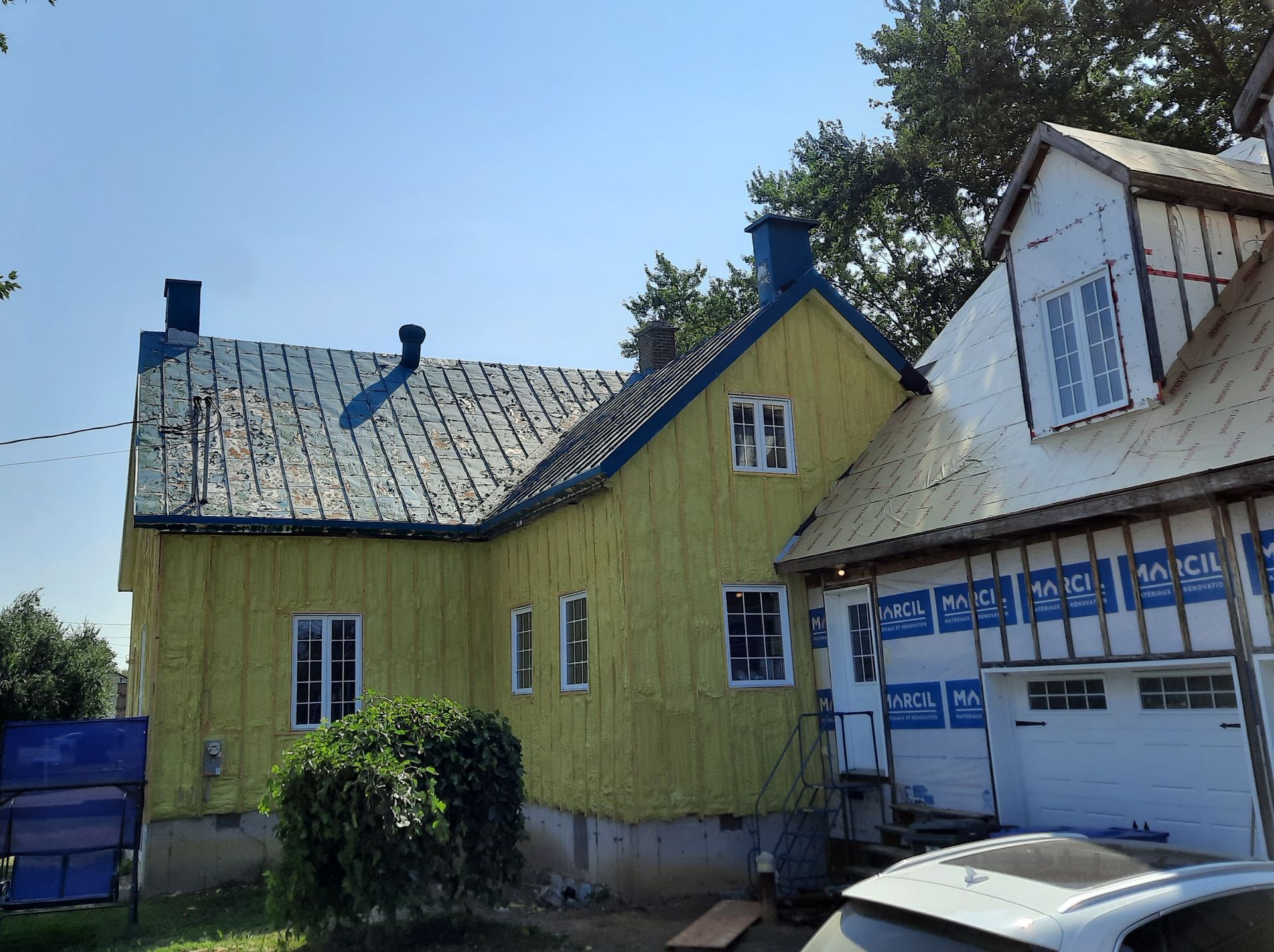 Maison jaune en construction ; bâche bleue, toit bleu, fenêtres blanches et bois apparents sont visibles.