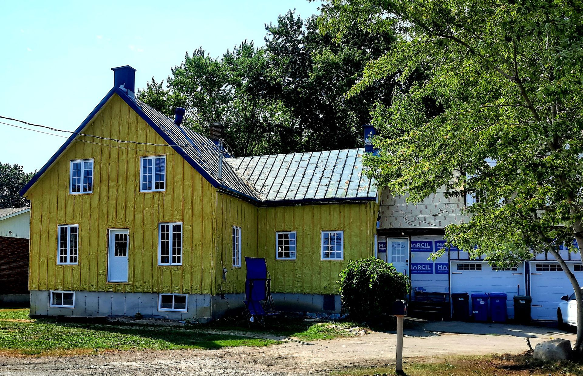 Maison jaune avec des bordures bleues, sur fond d'arbres verts, journée ensoleillée.