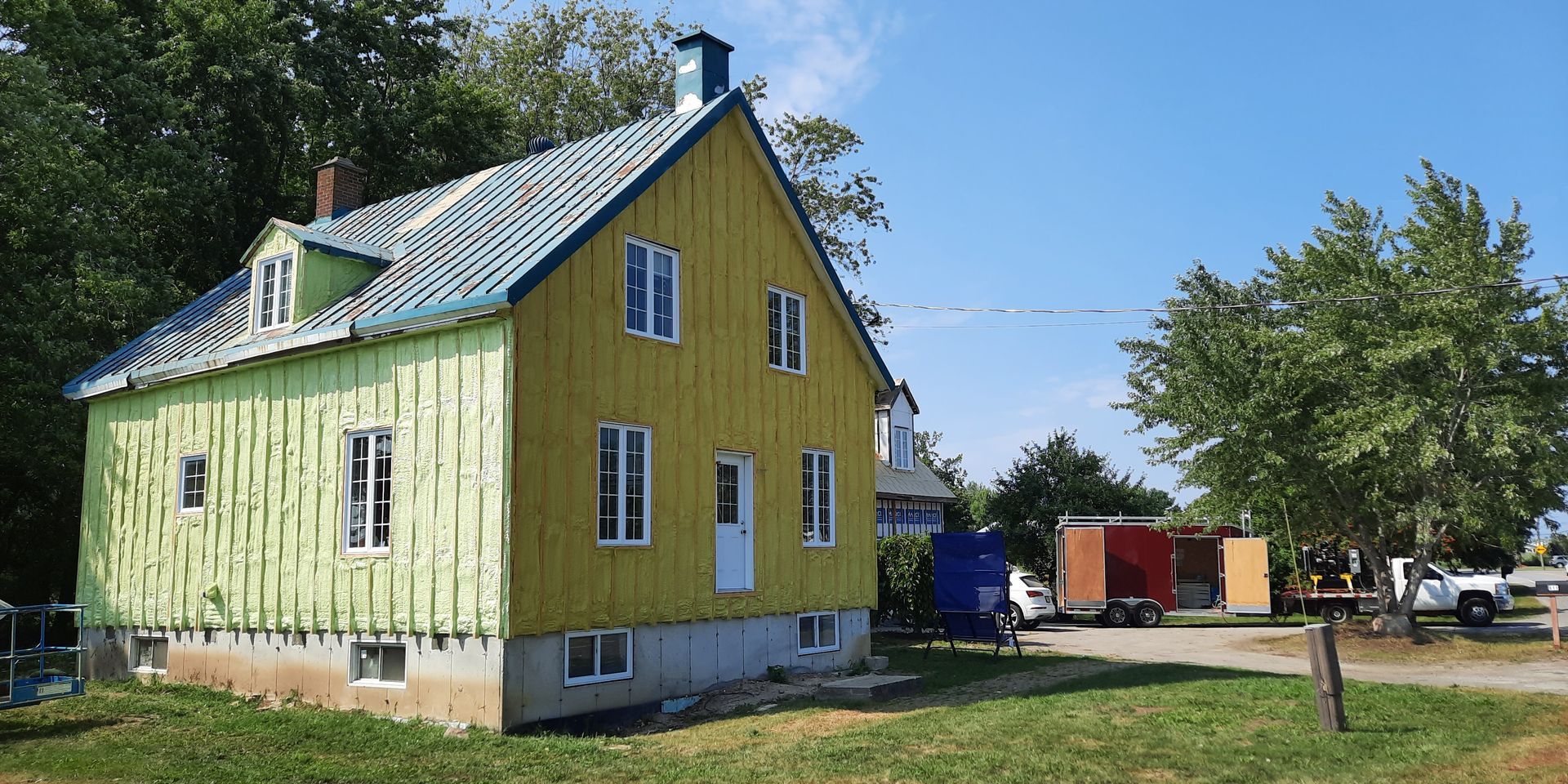 Maison jaune avec garniture bleue, caravanes garées et un arbre sous un ciel bleu.