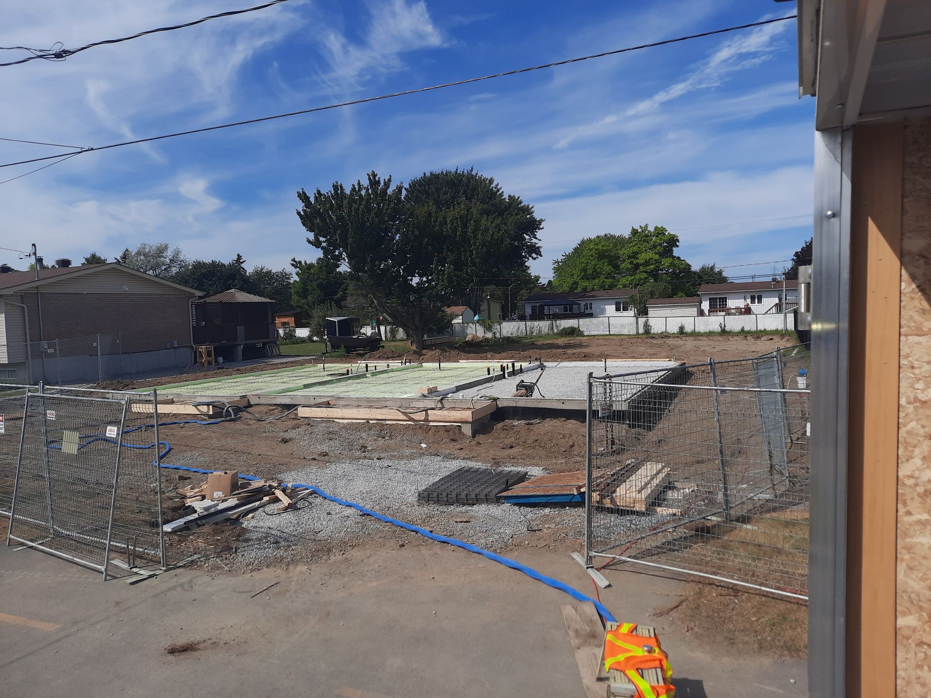 Chantier de construction avec fondation, gravier et un grand arbre sous un ciel bleu.