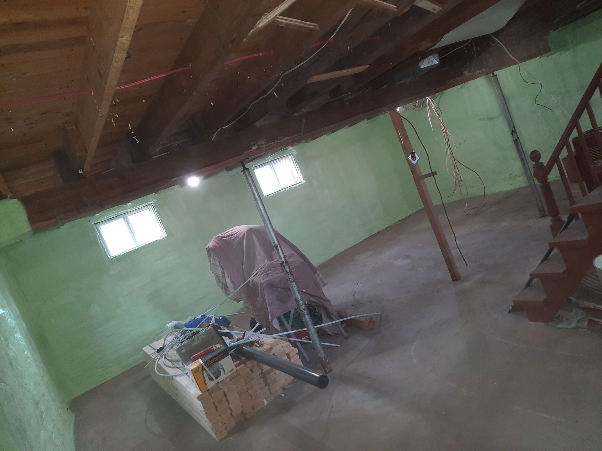 Sous-sol intérieur avec murs végétalisés, poutres en bois et sol en béton. On aperçoit un objet couvert et un escalier.